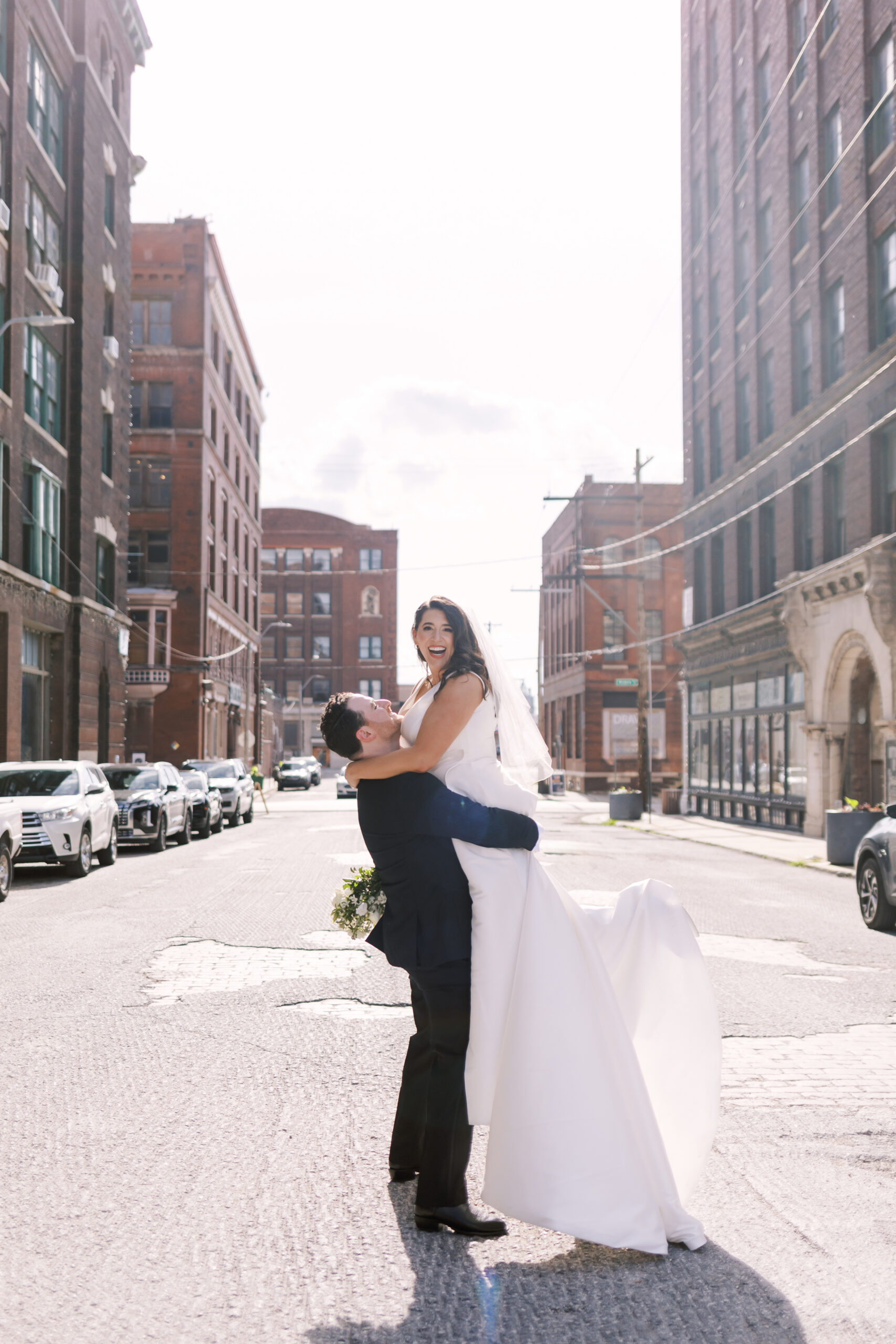 Groom lifting the bride in the middle of a city street as she smiles toward the camera.