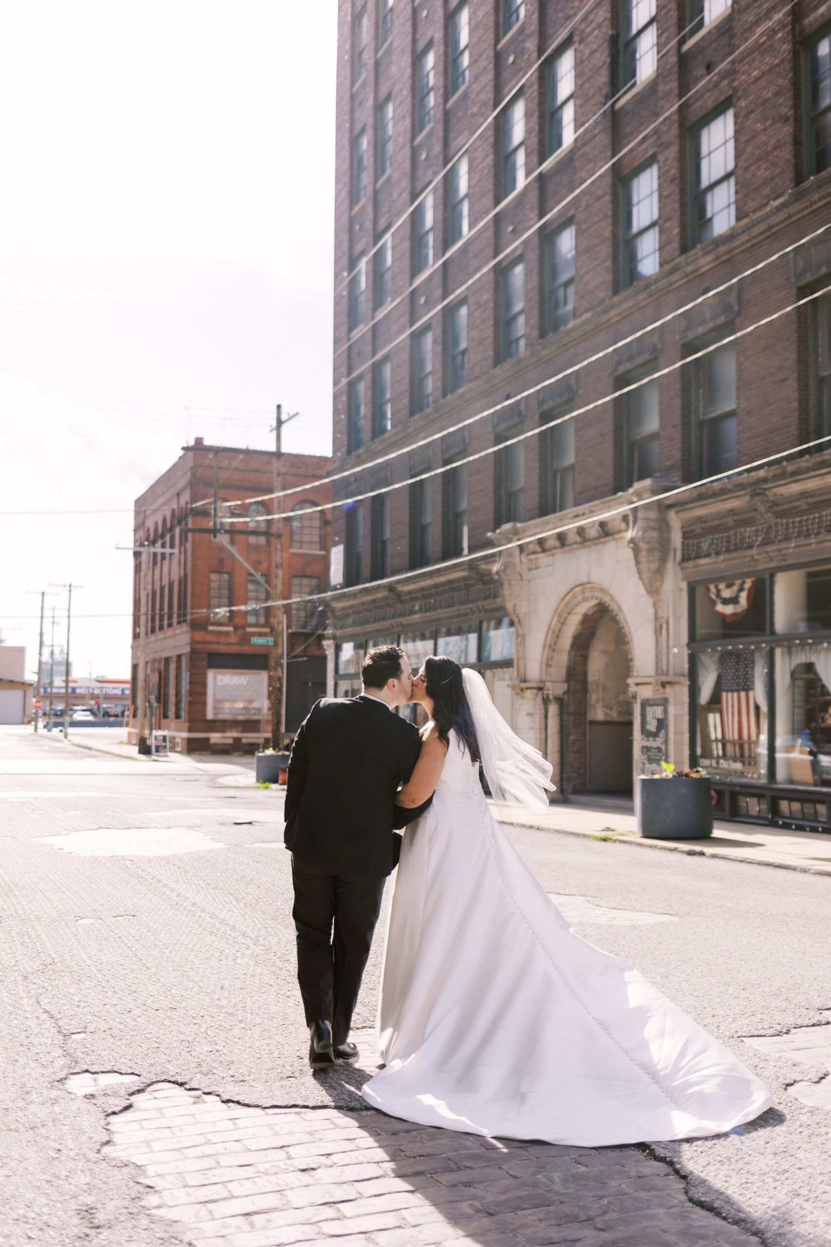 Bride and groom walking down a sunlit downtown street as they lean in for a kiss.