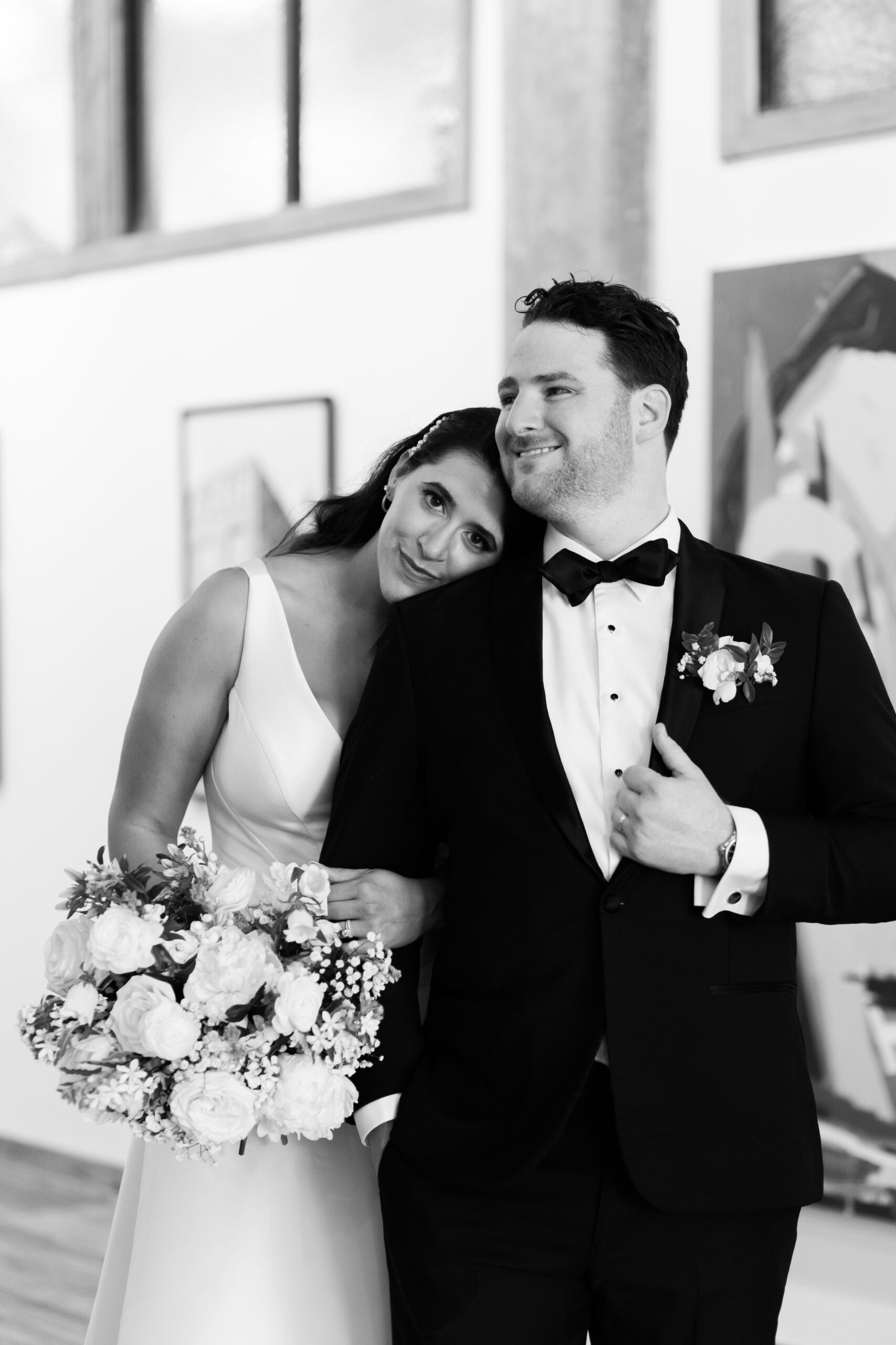Bride resting her head on the groom’s shoulder while holding a white bouquet indoors.