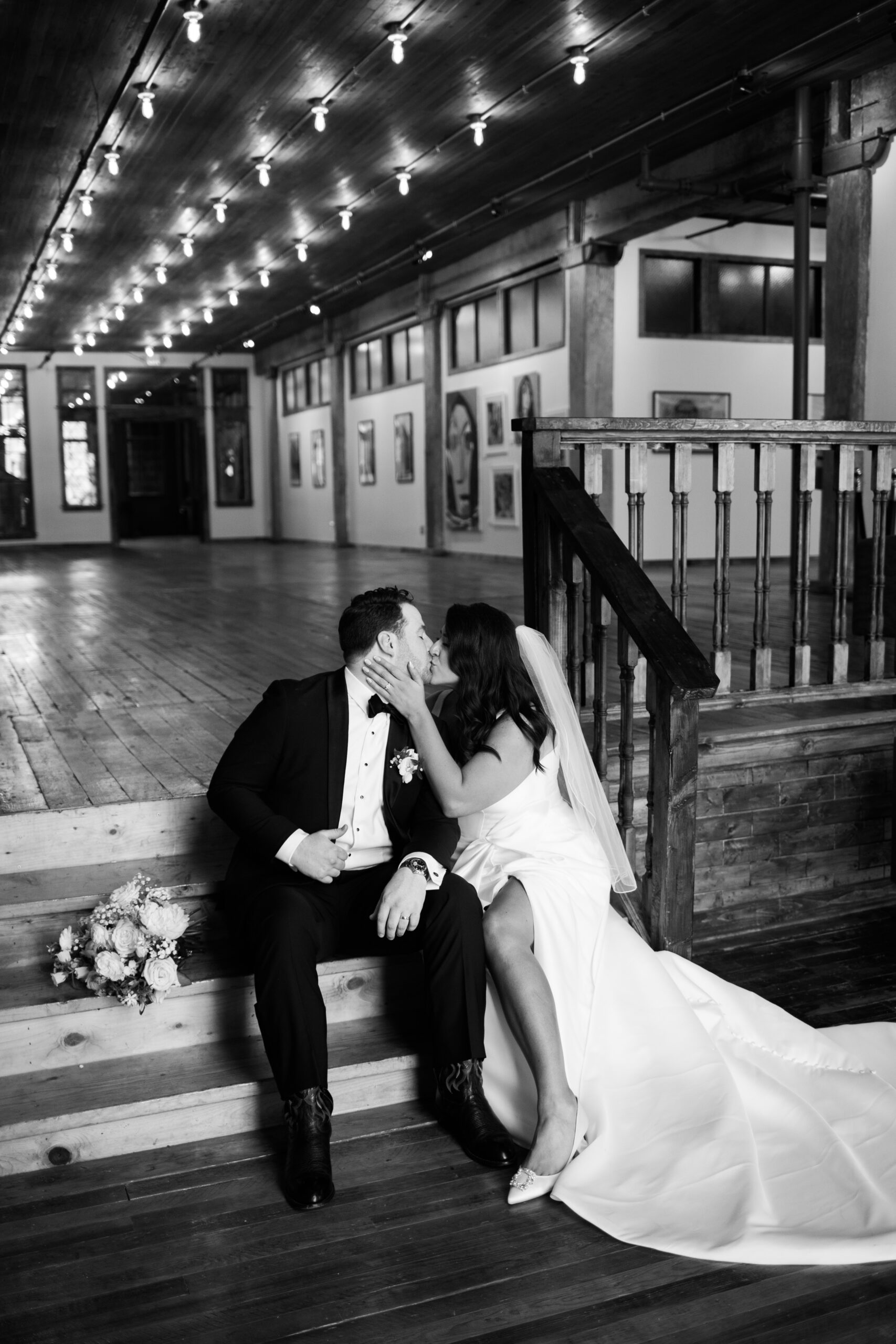 Bride and groom seated on wooden steps sharing a kiss beneath string lights in a loft venue.