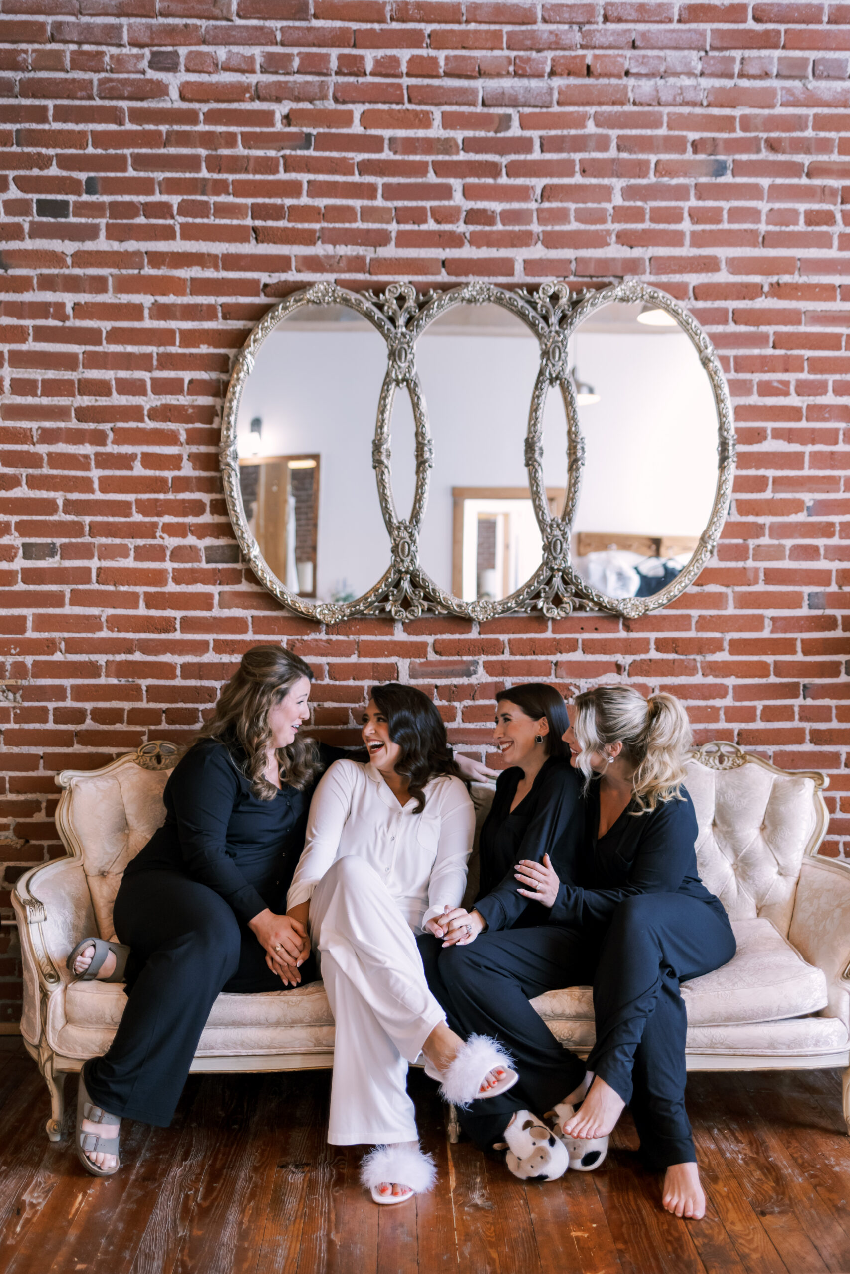 Bride sitting on a couch laughing with bridesmaids in matching black outfits before the ceremony.