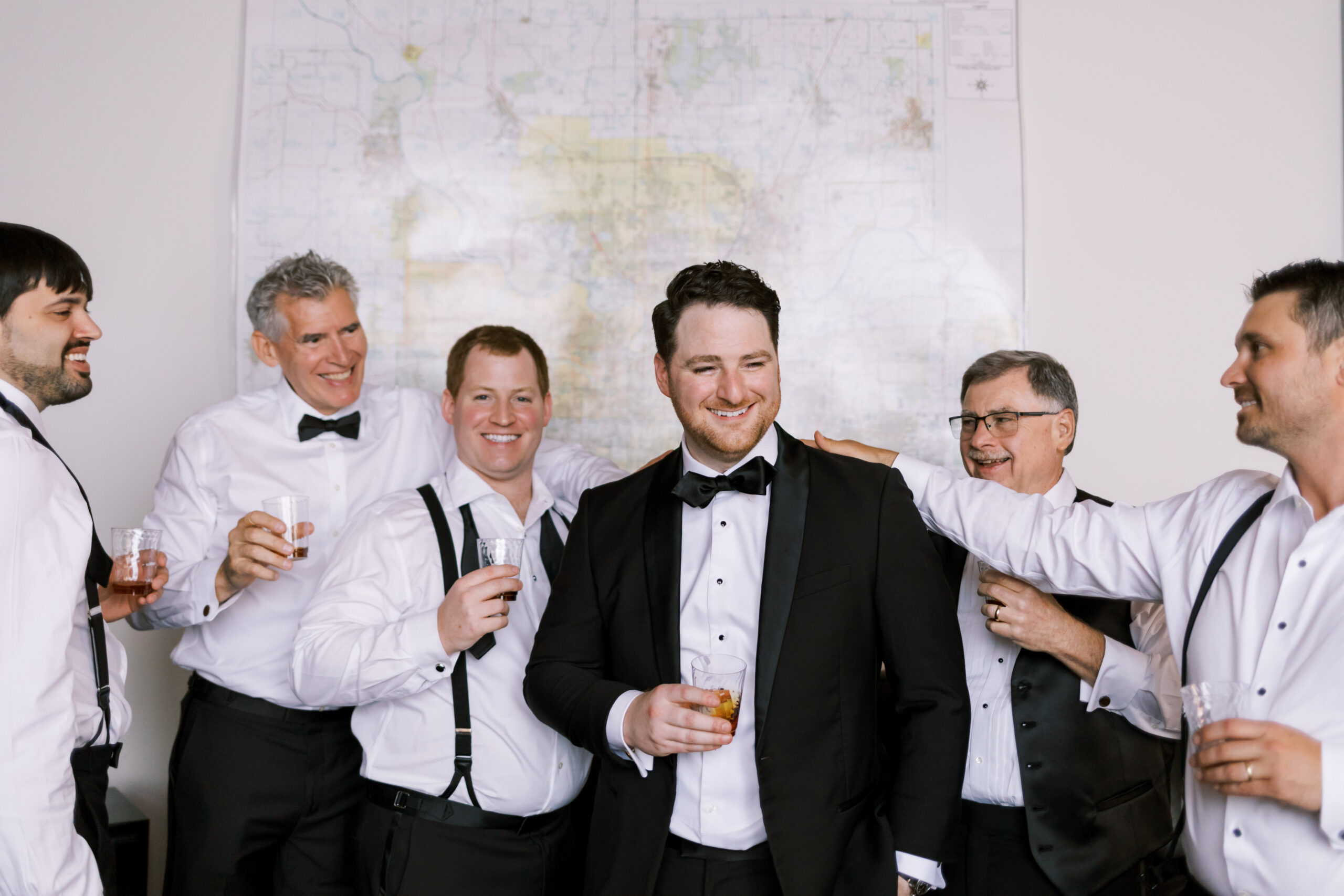 Groom in a tuxedo standing with groomsmen holding drinks during pre-ceremony preparations.