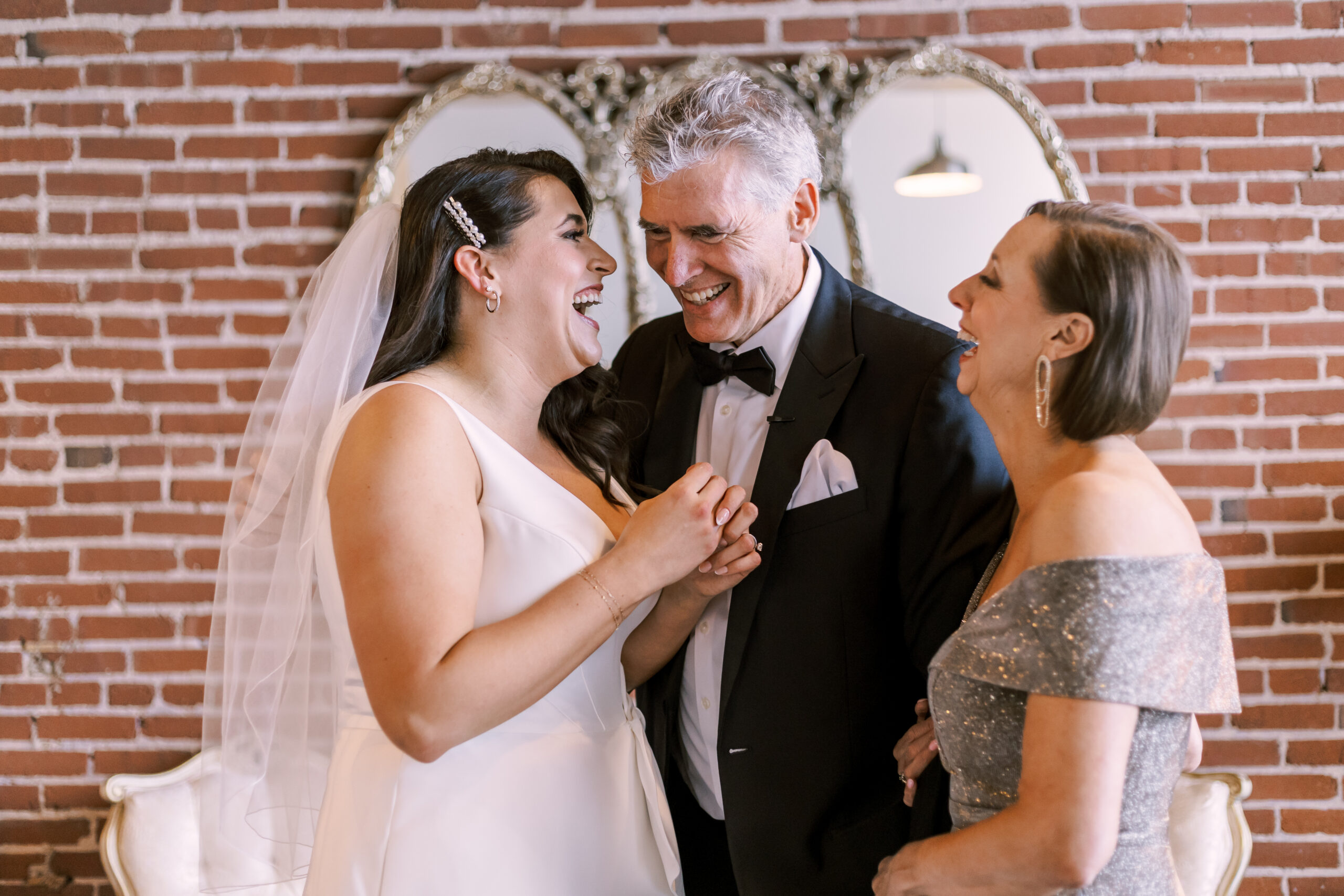 Bride standing with her parents laughing together in front of a brick wall and decorative mirror.