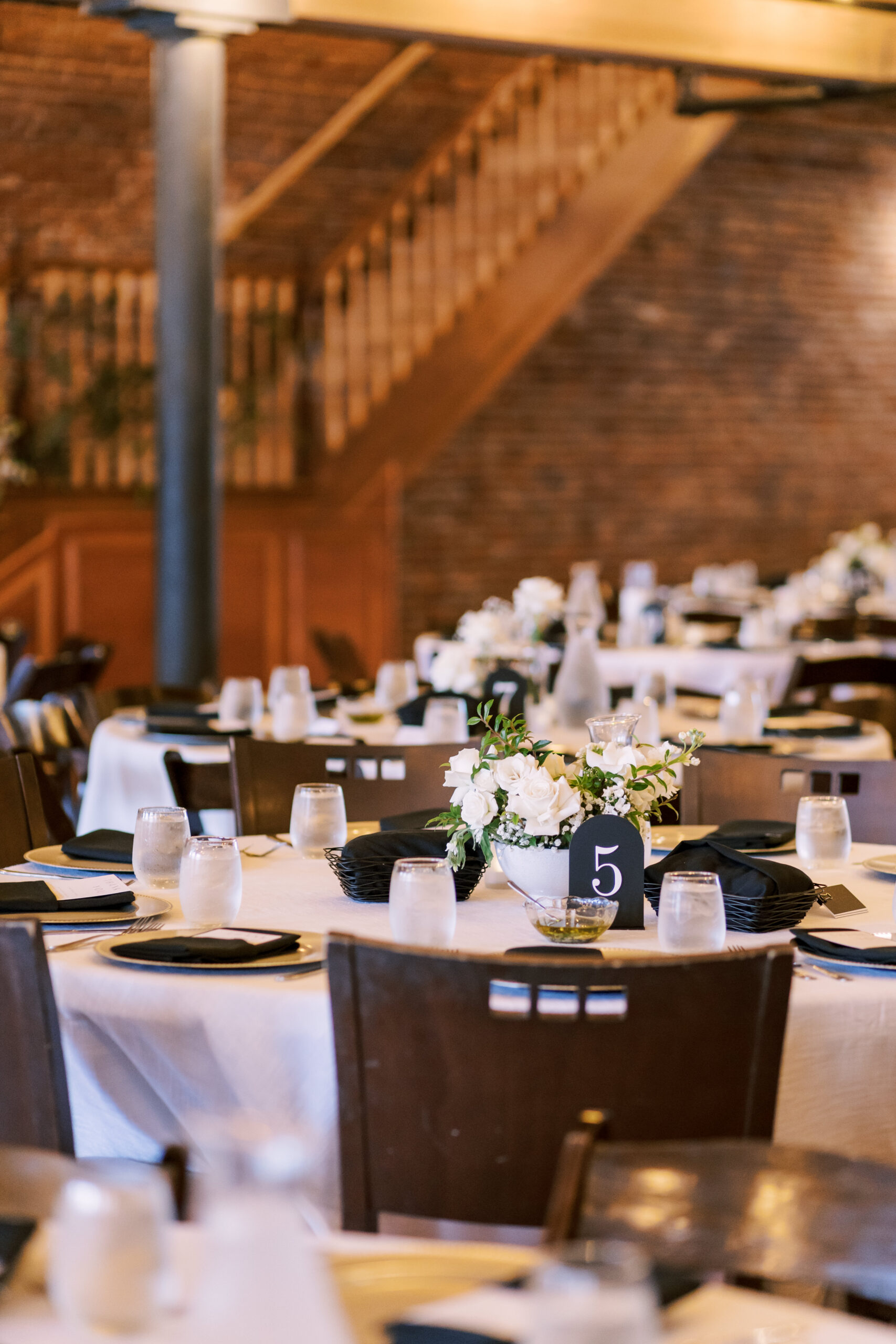 Round reception tables with white linens, black napkins, and floral centerpieces inside a brick loft wedding venue.