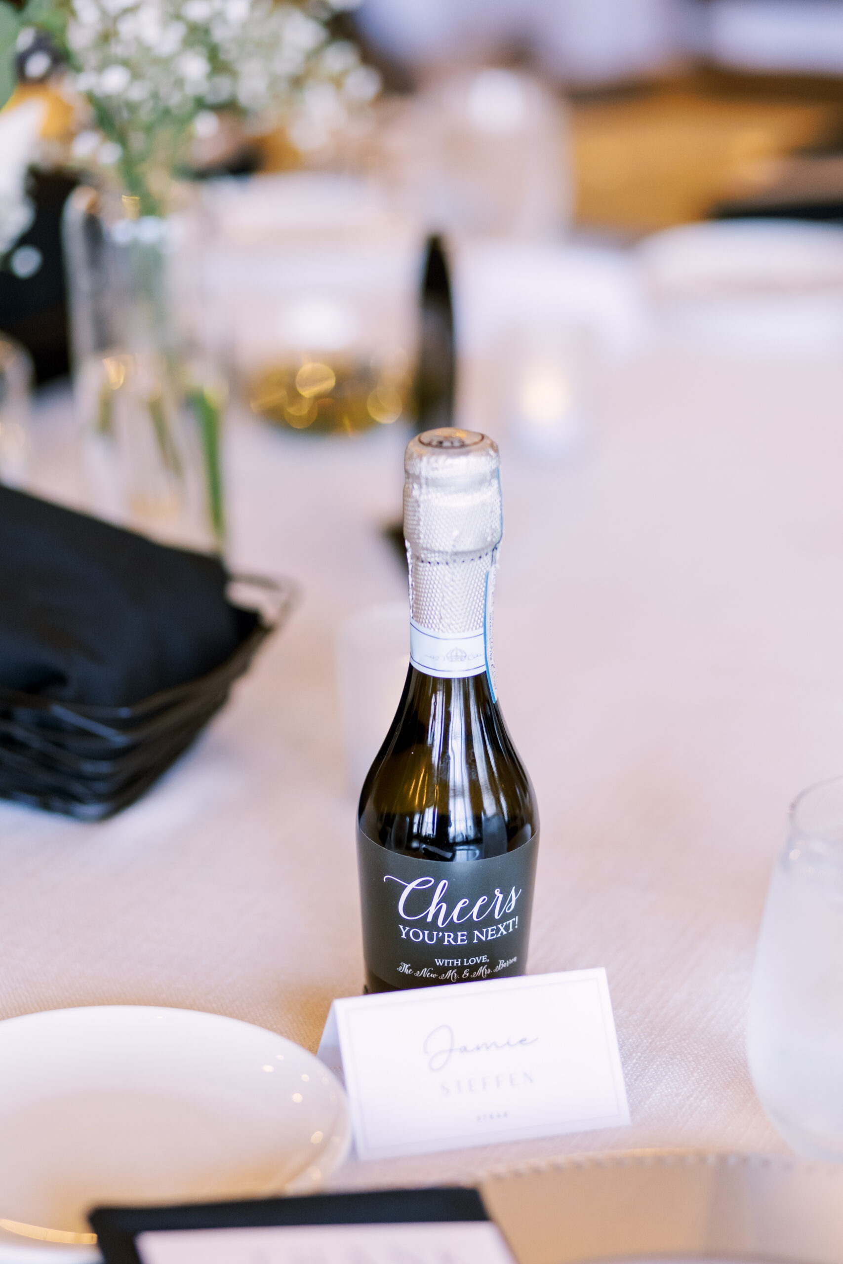 Mini champagne bottle wedding favor labeled “Cheers, you’re next” placed at a guest table setting.