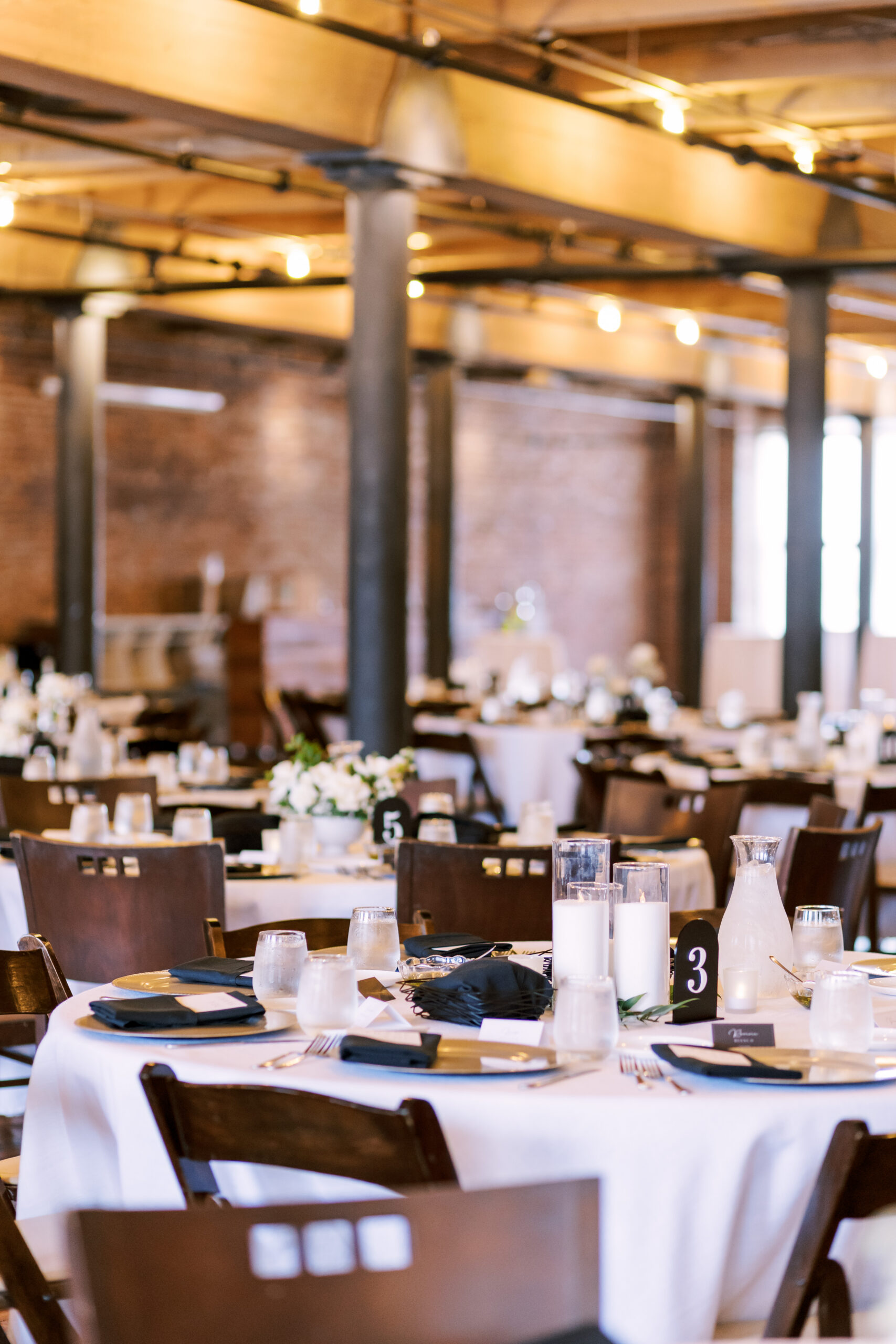 Wedding reception tables arranged throughout a brick loft venue with warm string lighting overhead.