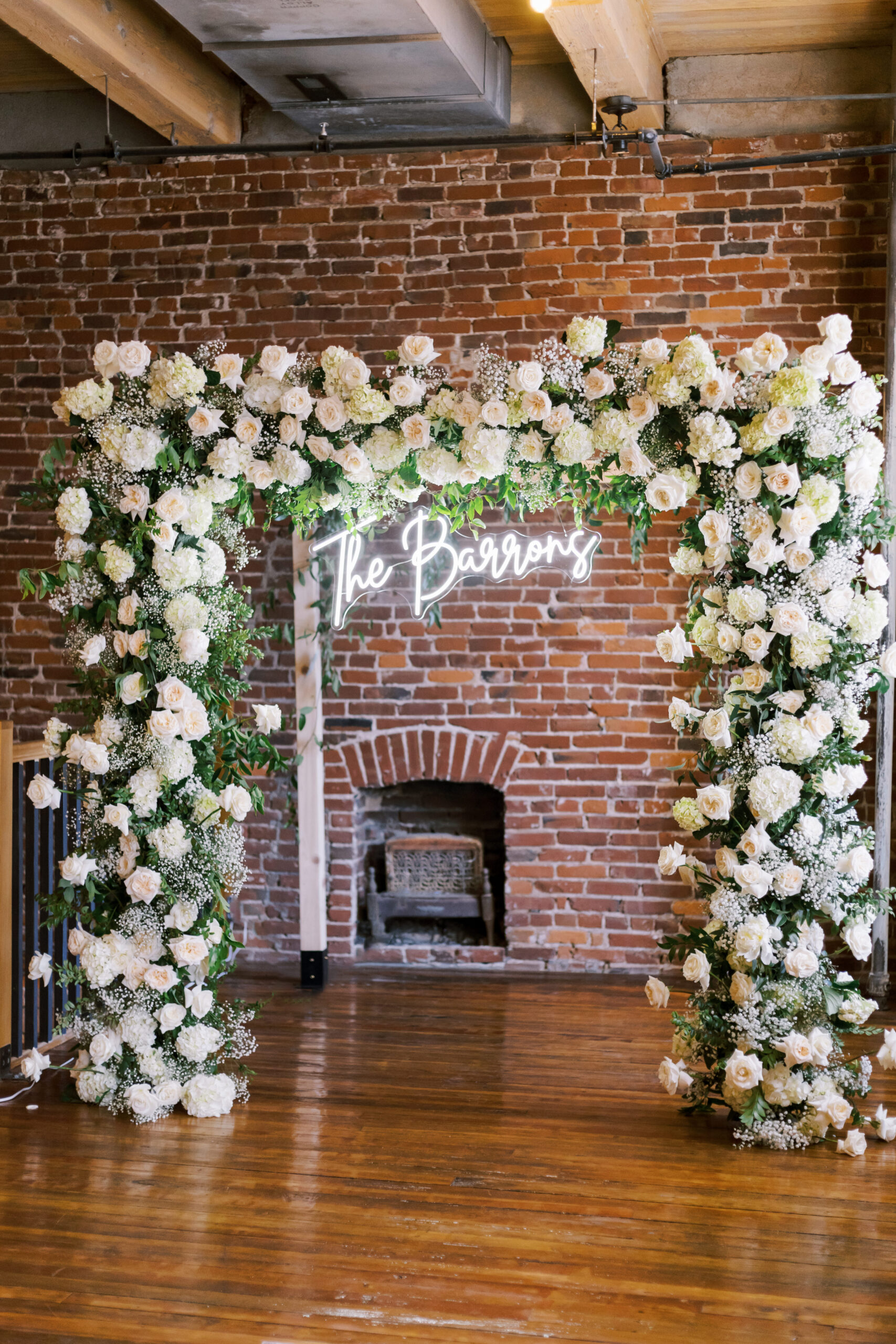 White rose and greenery ceremony arch with a neon last-name sign in front of a brick wall.