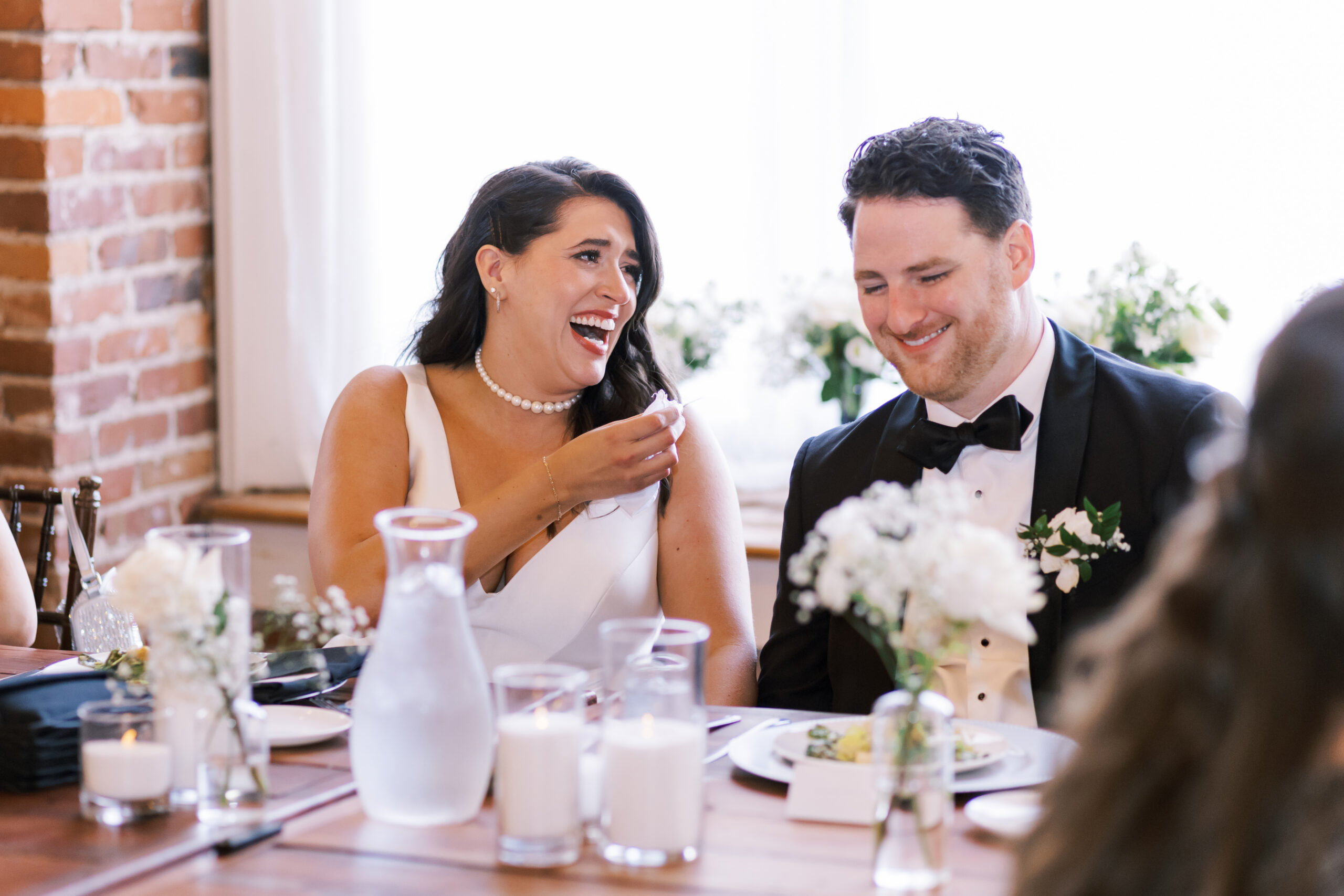 Bride laughing beside the groom while seated at their reception table.