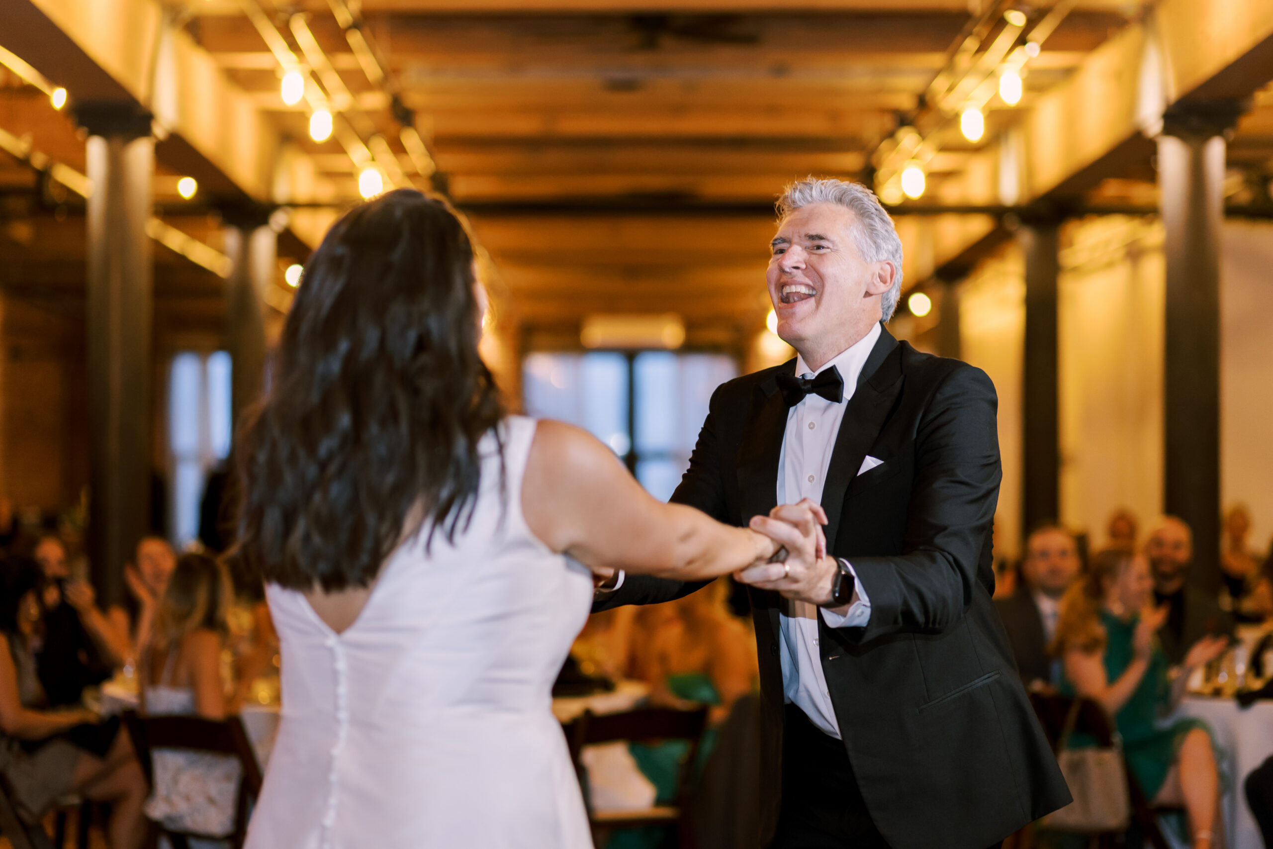 Bride dancing with an older man in a tuxedo during a reception dance under warm string lights.