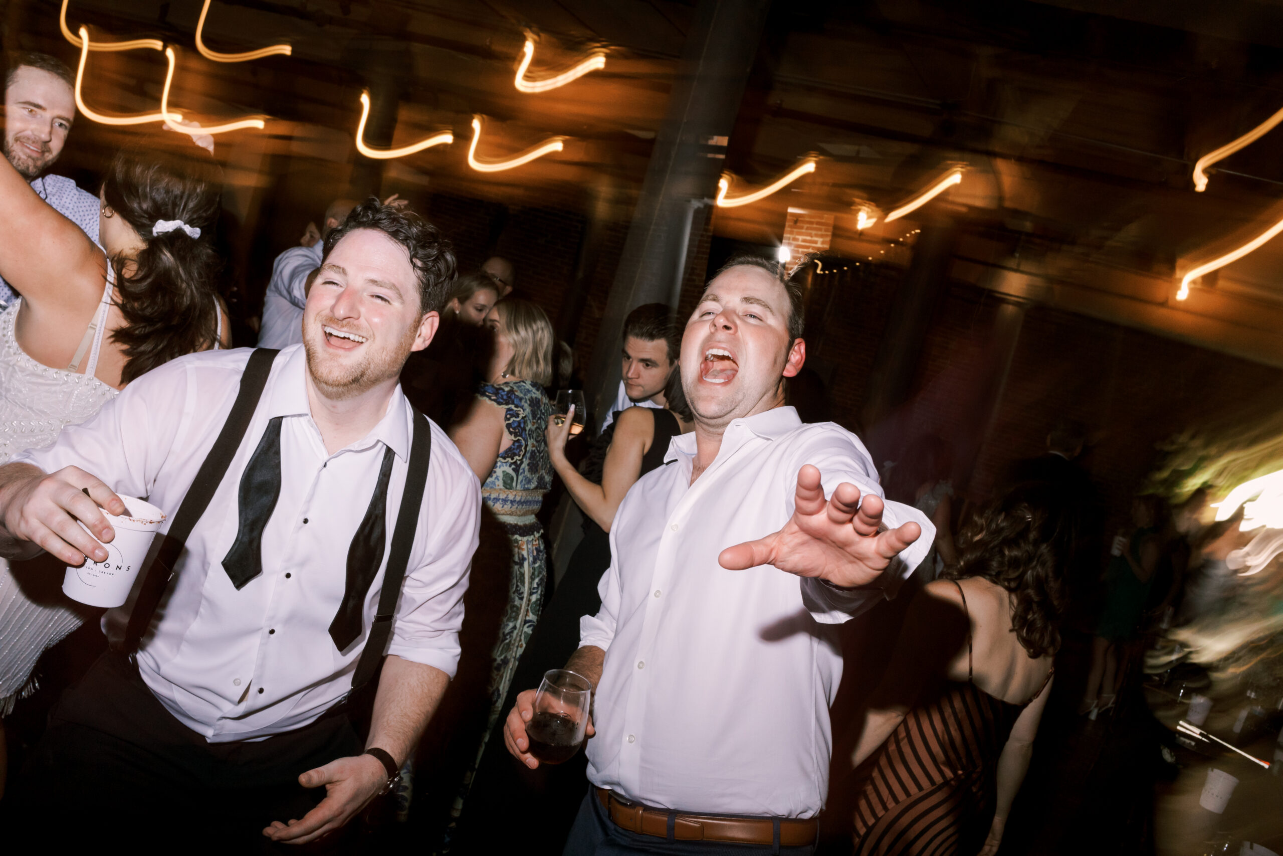 Two men in white dress shirts laughing and dancing energetically at a wedding reception, string lights blurred overhead