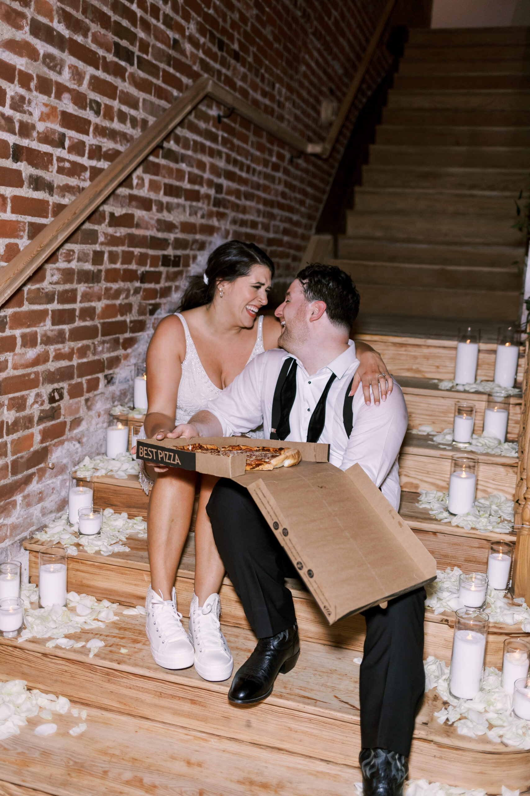 Bride and groom sitting on wooden steps with candles and rose petals while sharing late-night pizza.