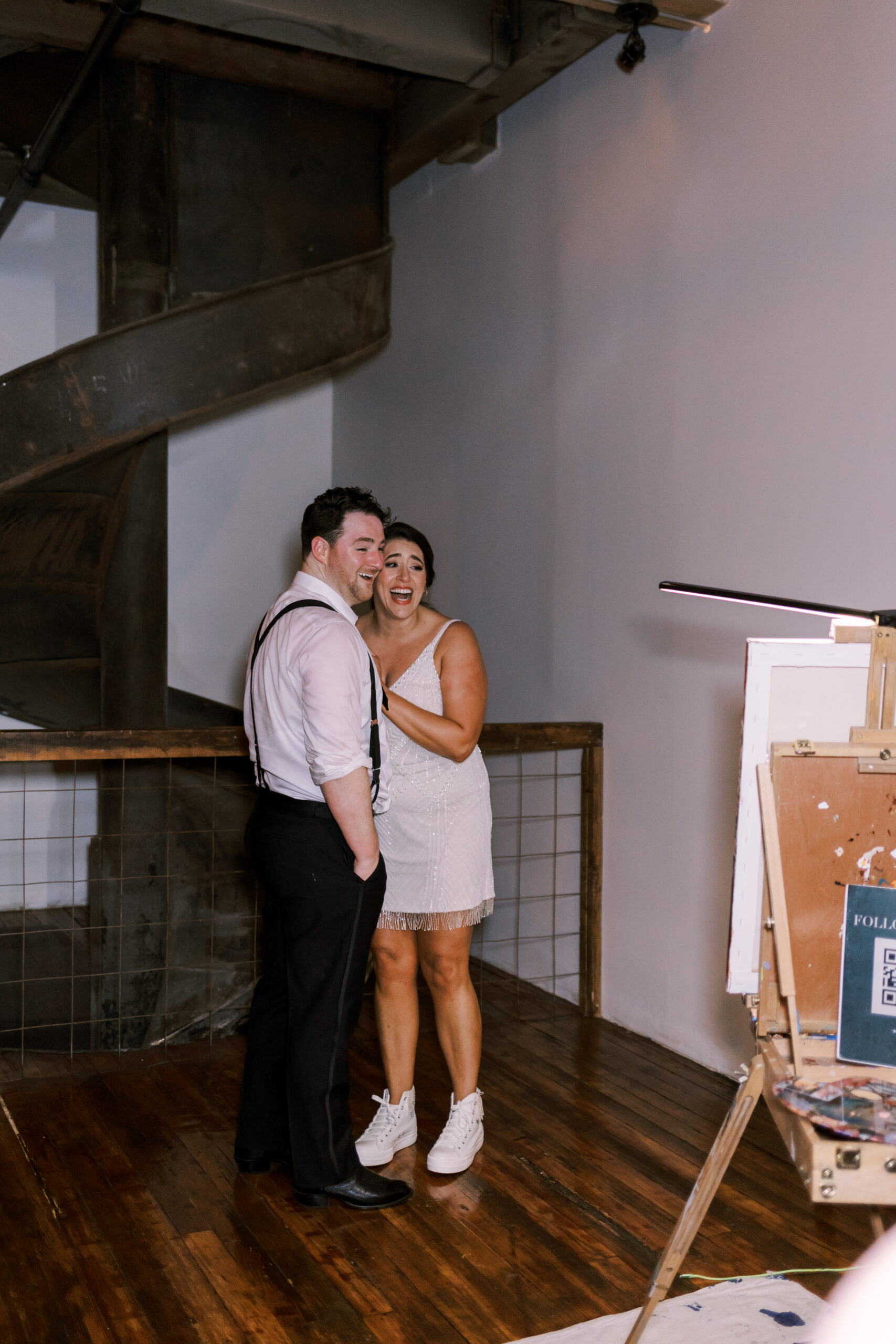Bride and groom laughing together near a spiral staircase beside a live wedding painter's easel