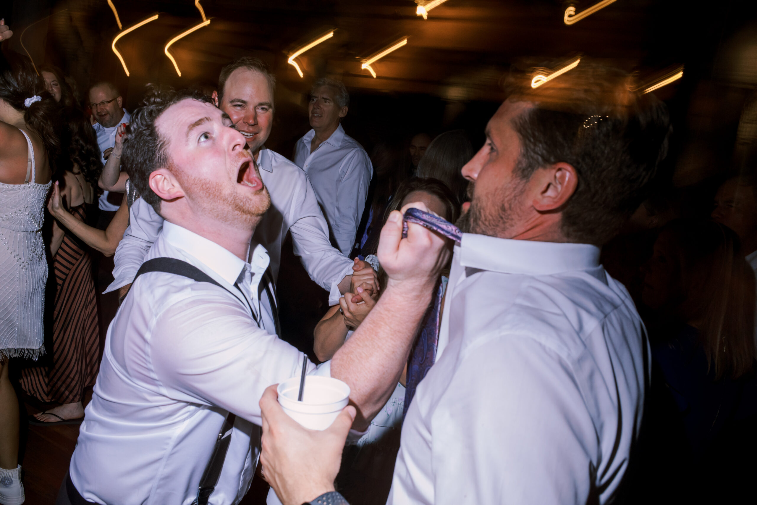 Man in suspenders reacting with exaggerated surprise as another man grabs his tie on the dance floor