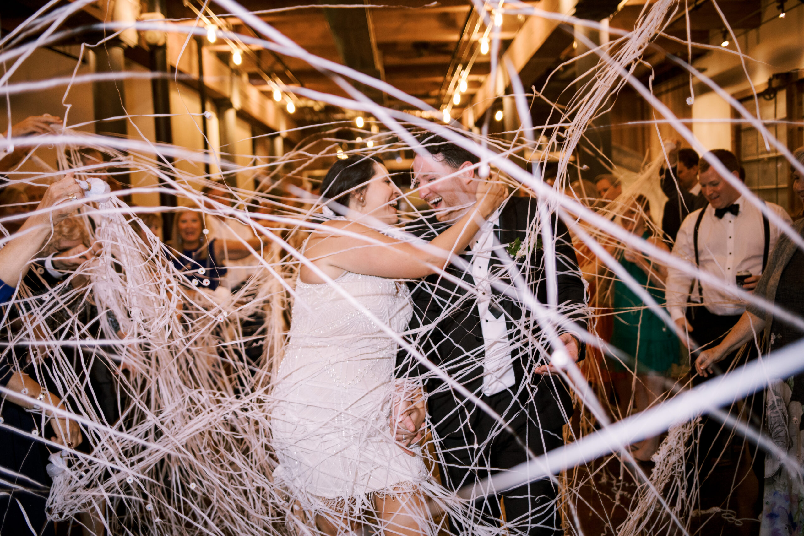 Bride and groom laughing together surrounded by streams of white confetti cannon ribbons on the dance floor
