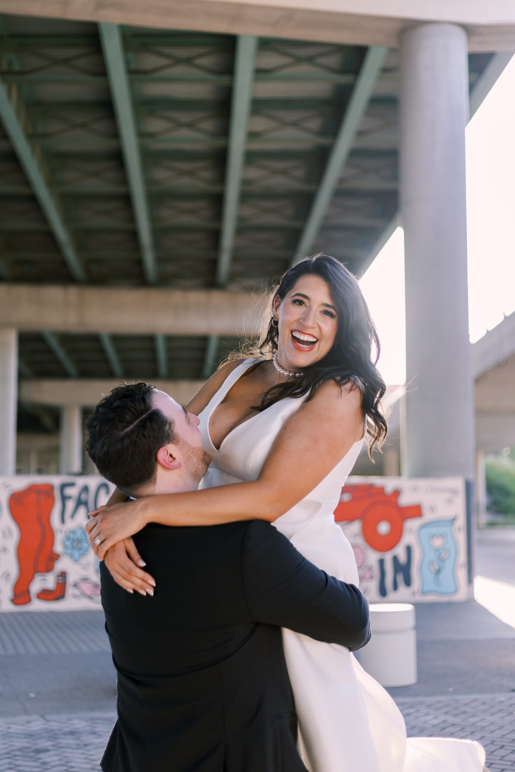Groom lifting and spinning a laughing bride in a white gown beneath an urban overpass with colorful graffiti murals