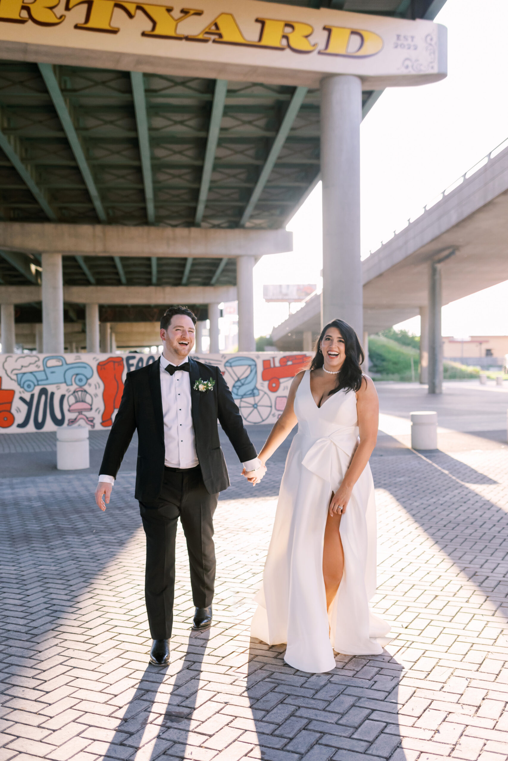 Bride and groom walking hand-in-hand and laughing beneath an overpass, colorful street murals visible behind them