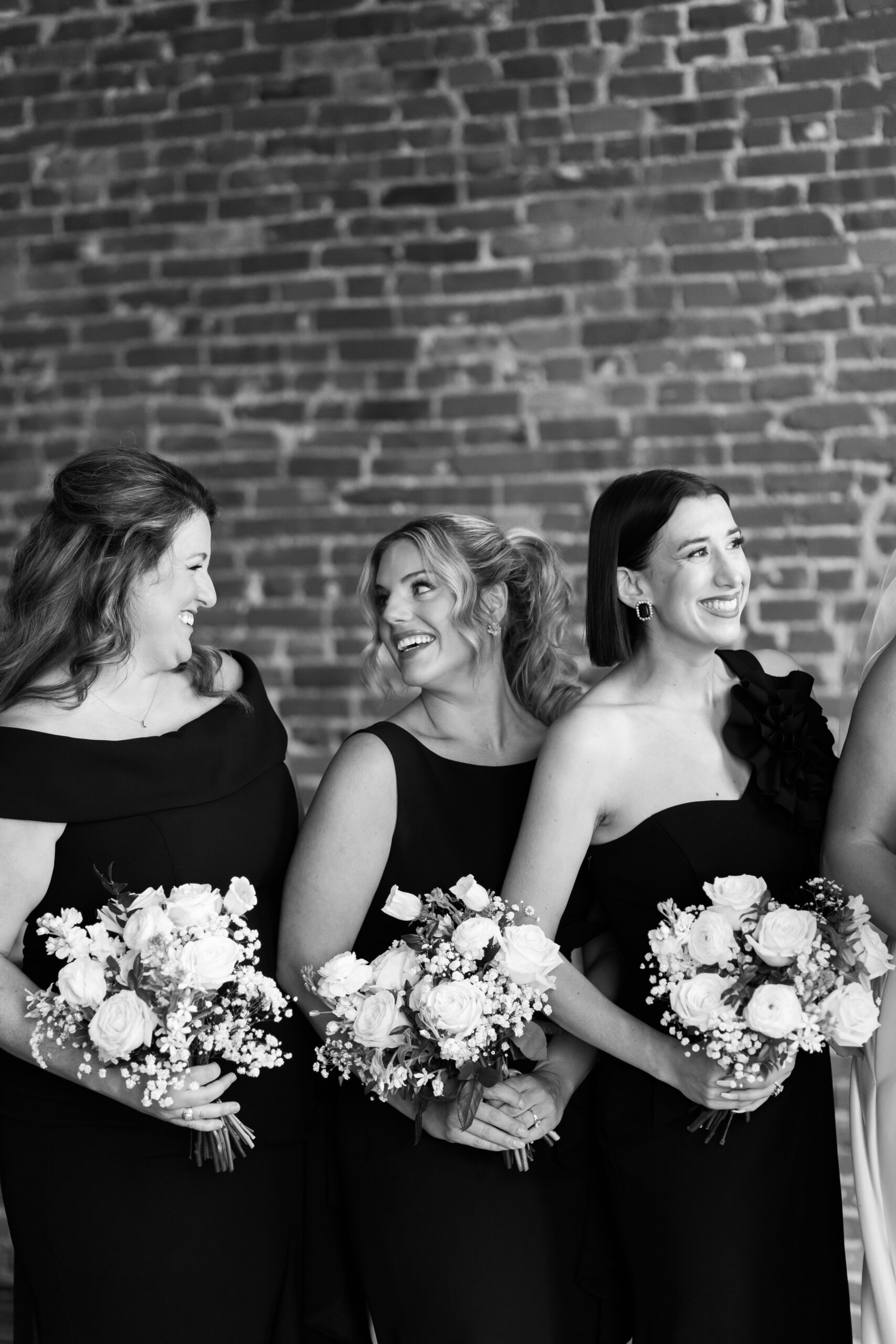 Black and white close-up of three bridesmaids in black gowns laughing together while holding rose bouquets against a brick wall