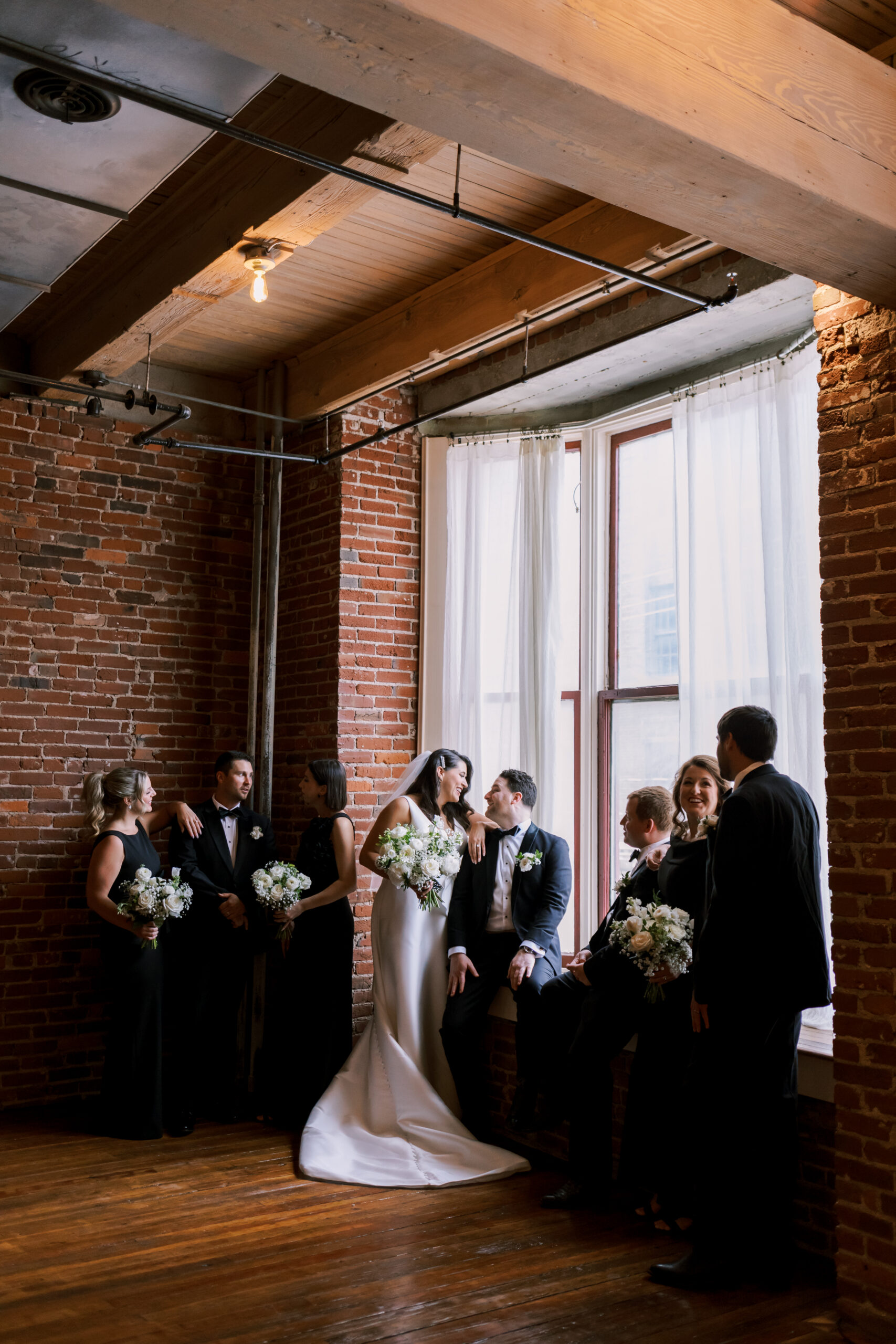 Wedding party of eight gathered around a large arched window in a brick-walled industrial venue, bride and groom at center