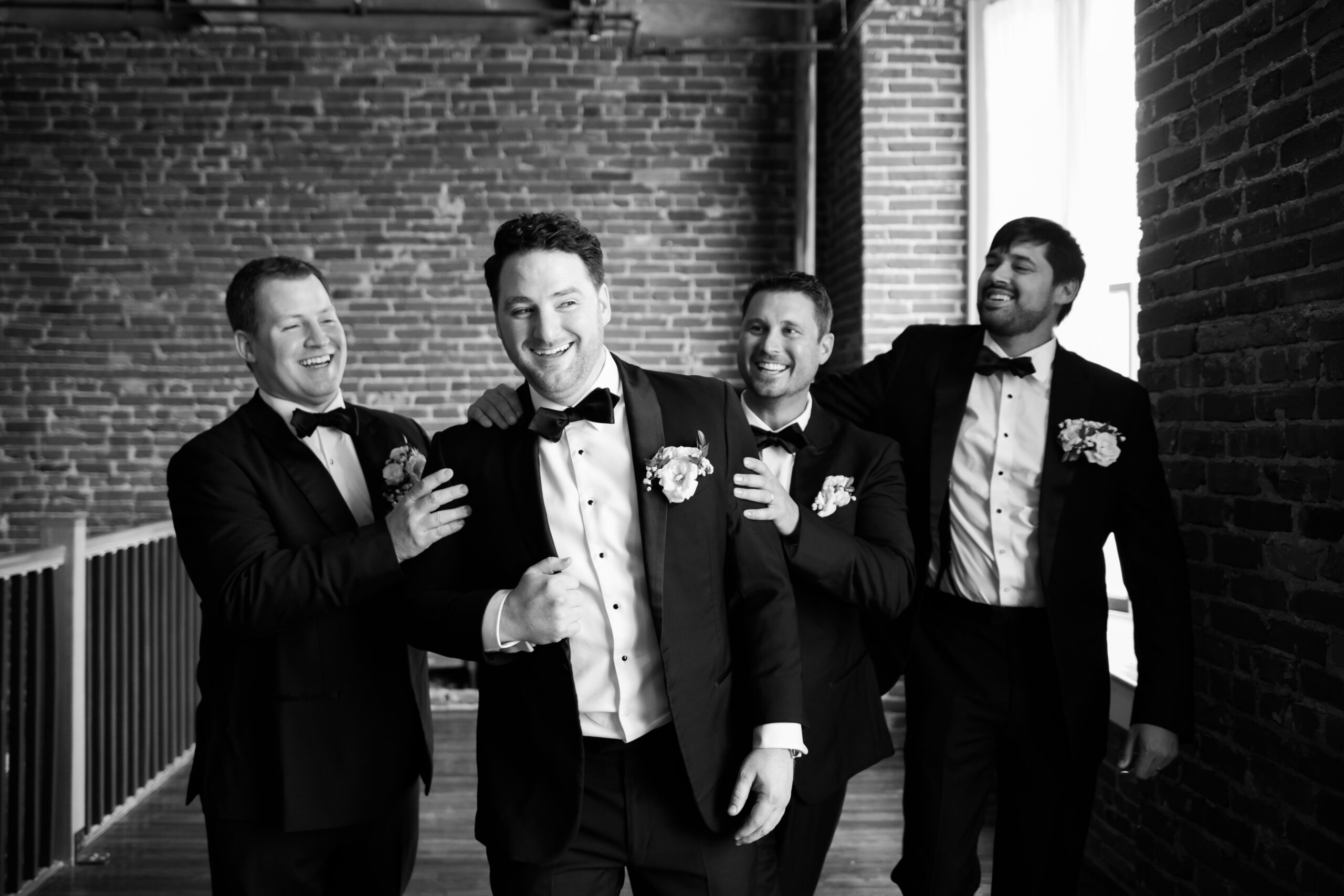 Black and white photo of the groom and three groomsmen in tuxedos laughing together against a brick wall