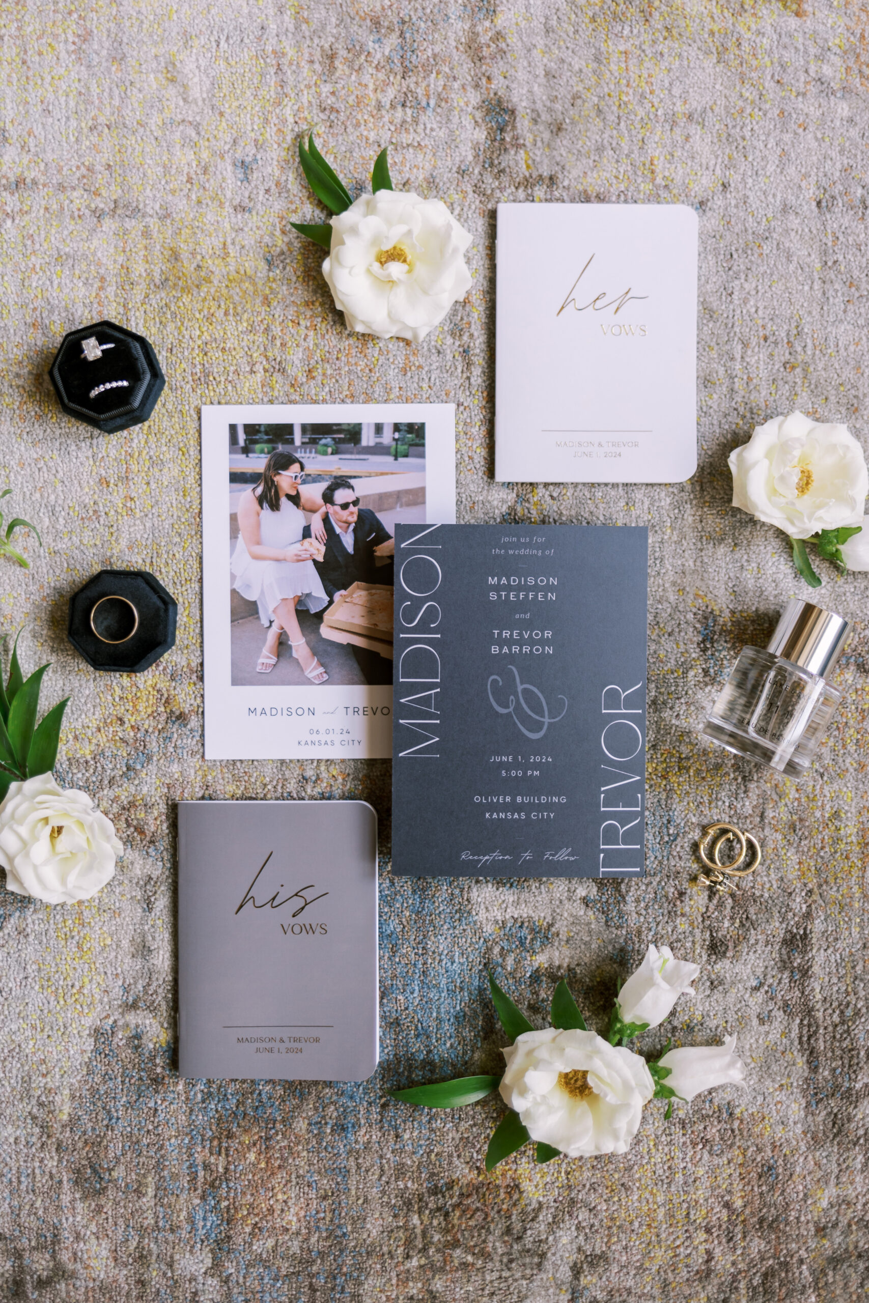Elegant wedding invitation suite with vow books, rings, perfume, and white flowers arranged on a textured rug.