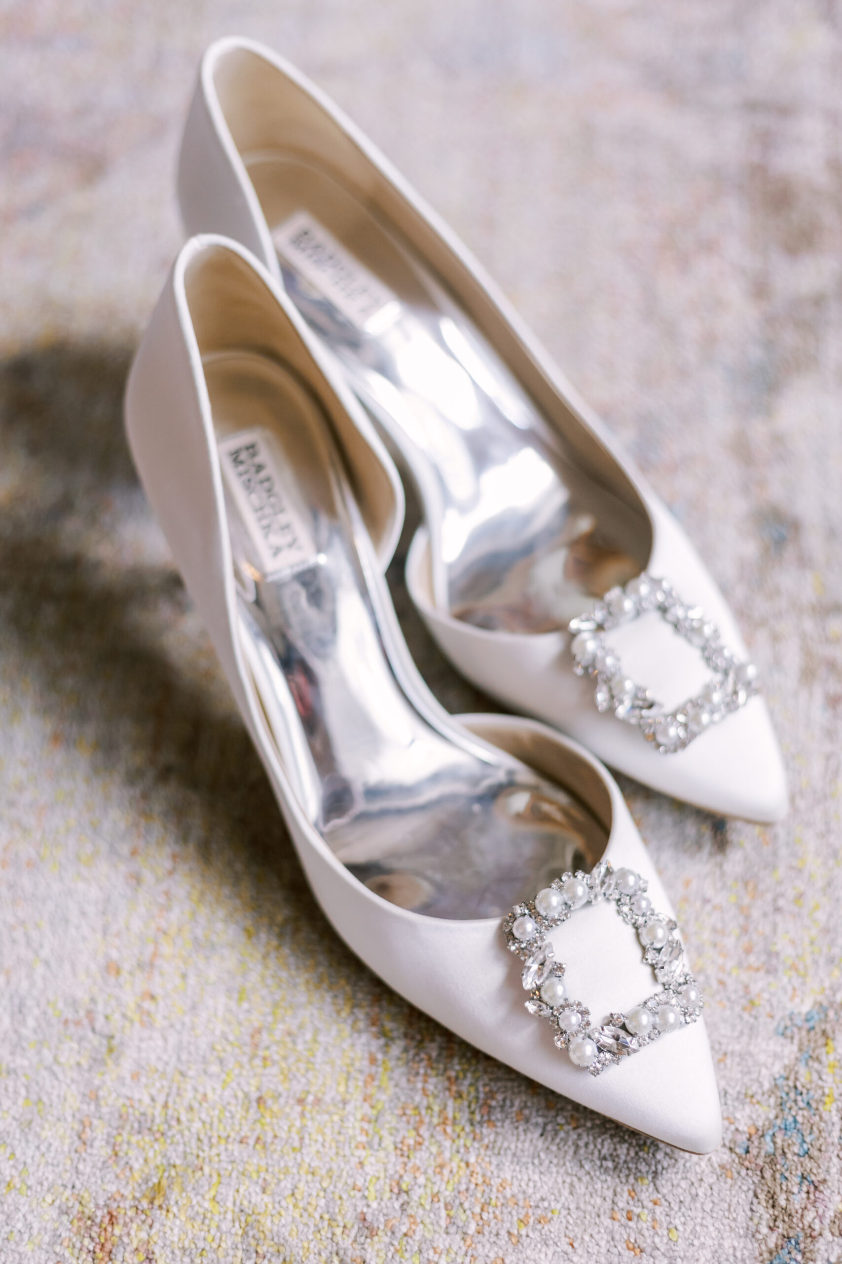 White pointed-toe bridal heels with jeweled buckle detail resting on a soft textured rug.