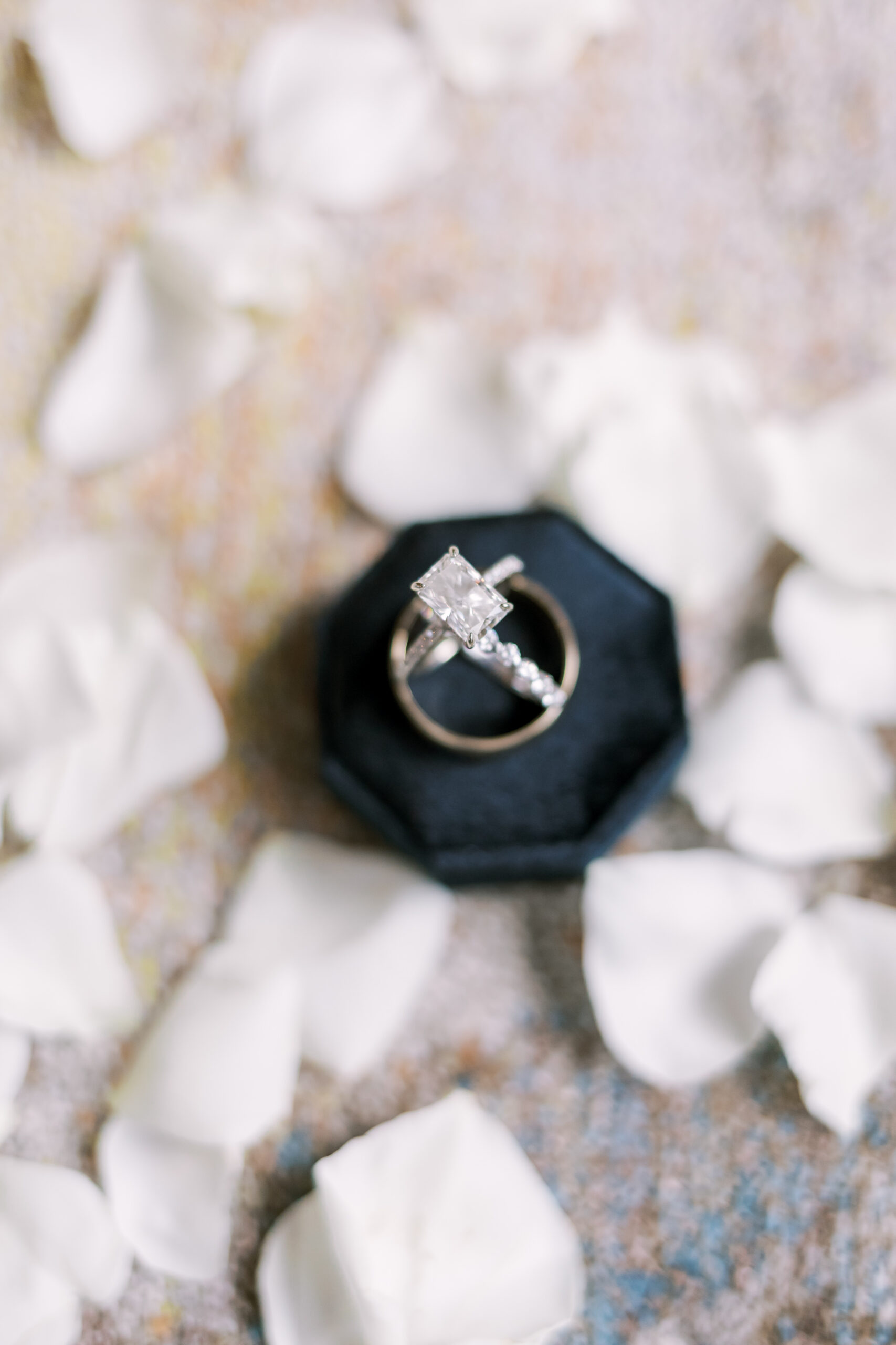 Emerald-cut engagement ring and diamond band in a black ring box surrounded by scattered white flower petals.