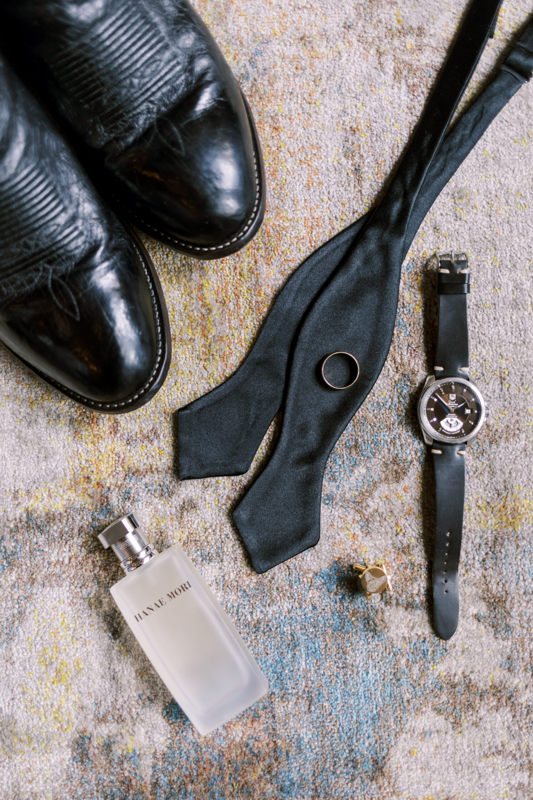 Black dress shoes, bow tie, watch, cufflinks, wedding band, and cologne arranged on a textured rug.