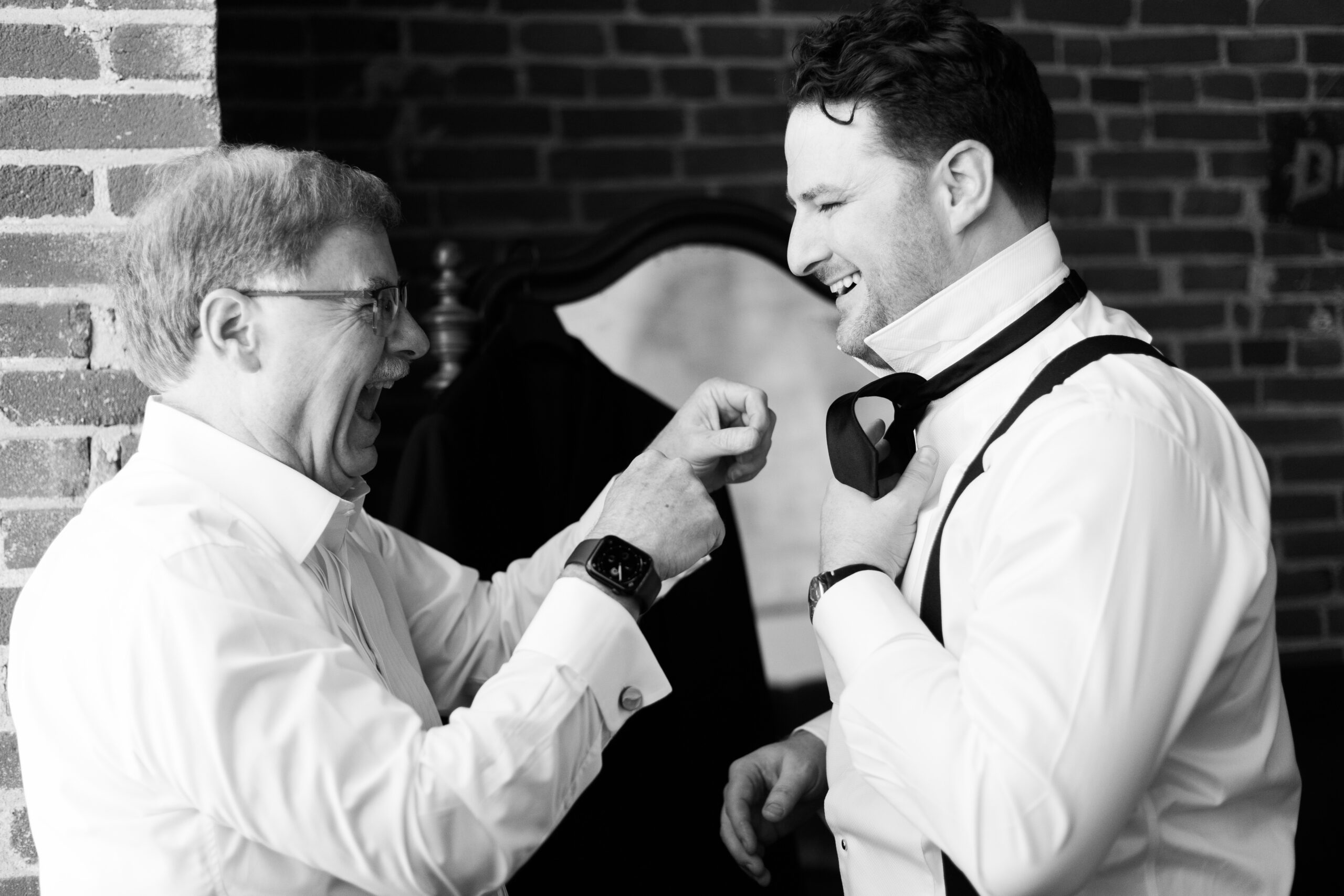 Older man adjusting a groom’s bow tie as they smile together in a brick-walled room.