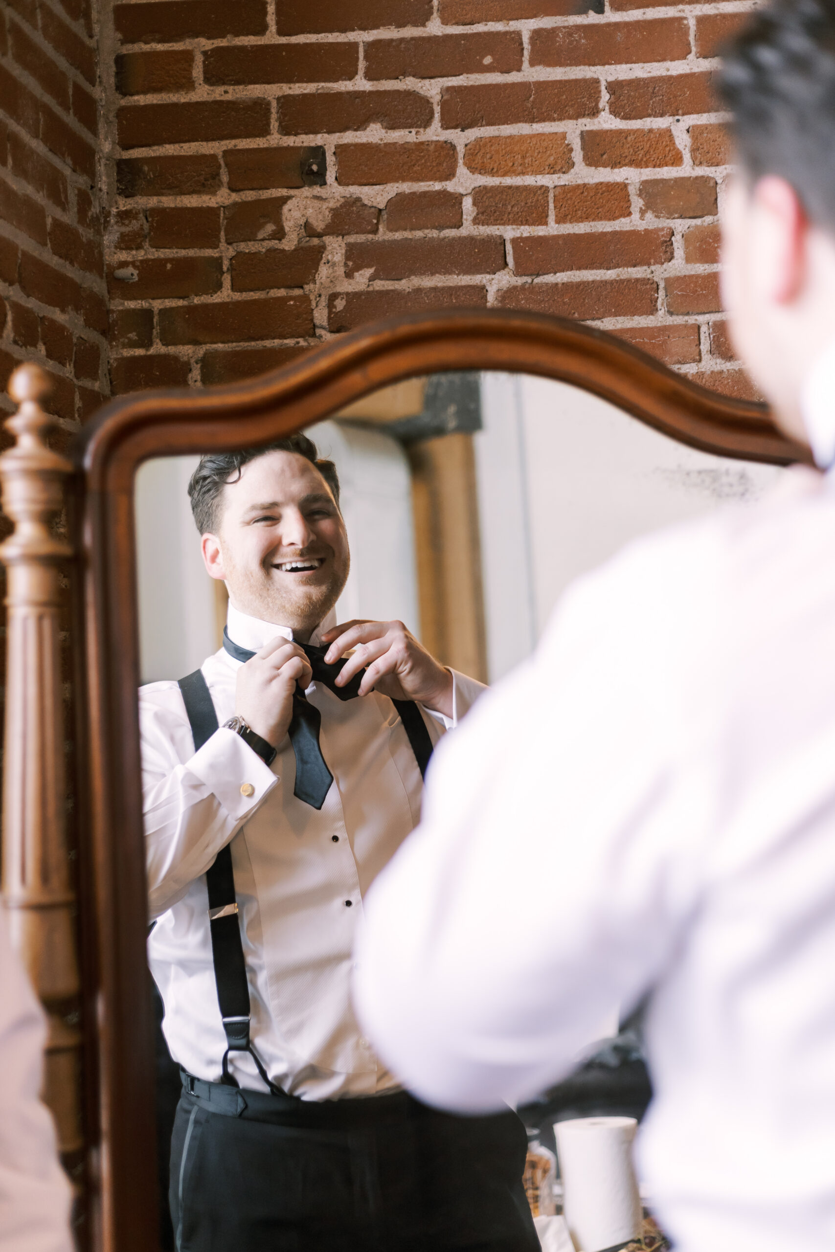 Groom smiling while adjusting his black bow tie in a mirror, reflected against an exposed brick wall.