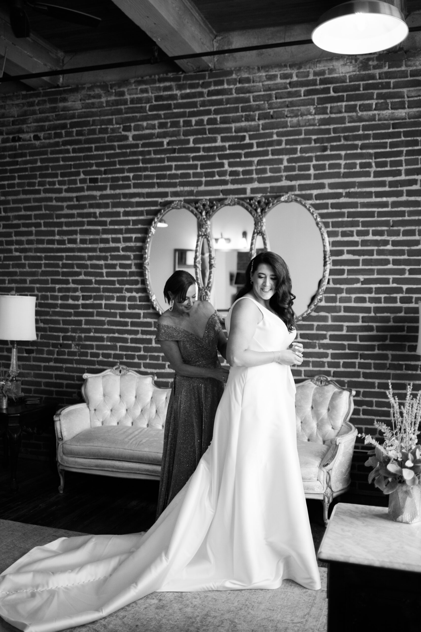 Bride standing in her wedding gown while another woman fastens the back in a brick loft room.