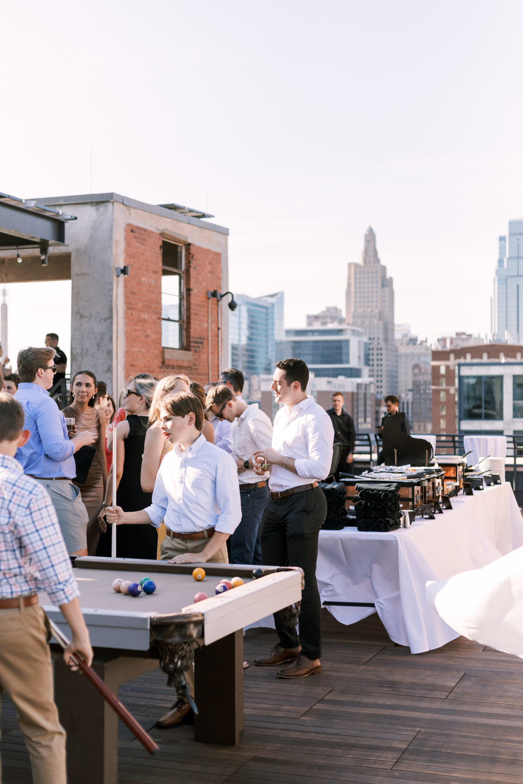 Guests gathered on a rooftop patio playing pool and chatting beside a buffet table with city skyline views.