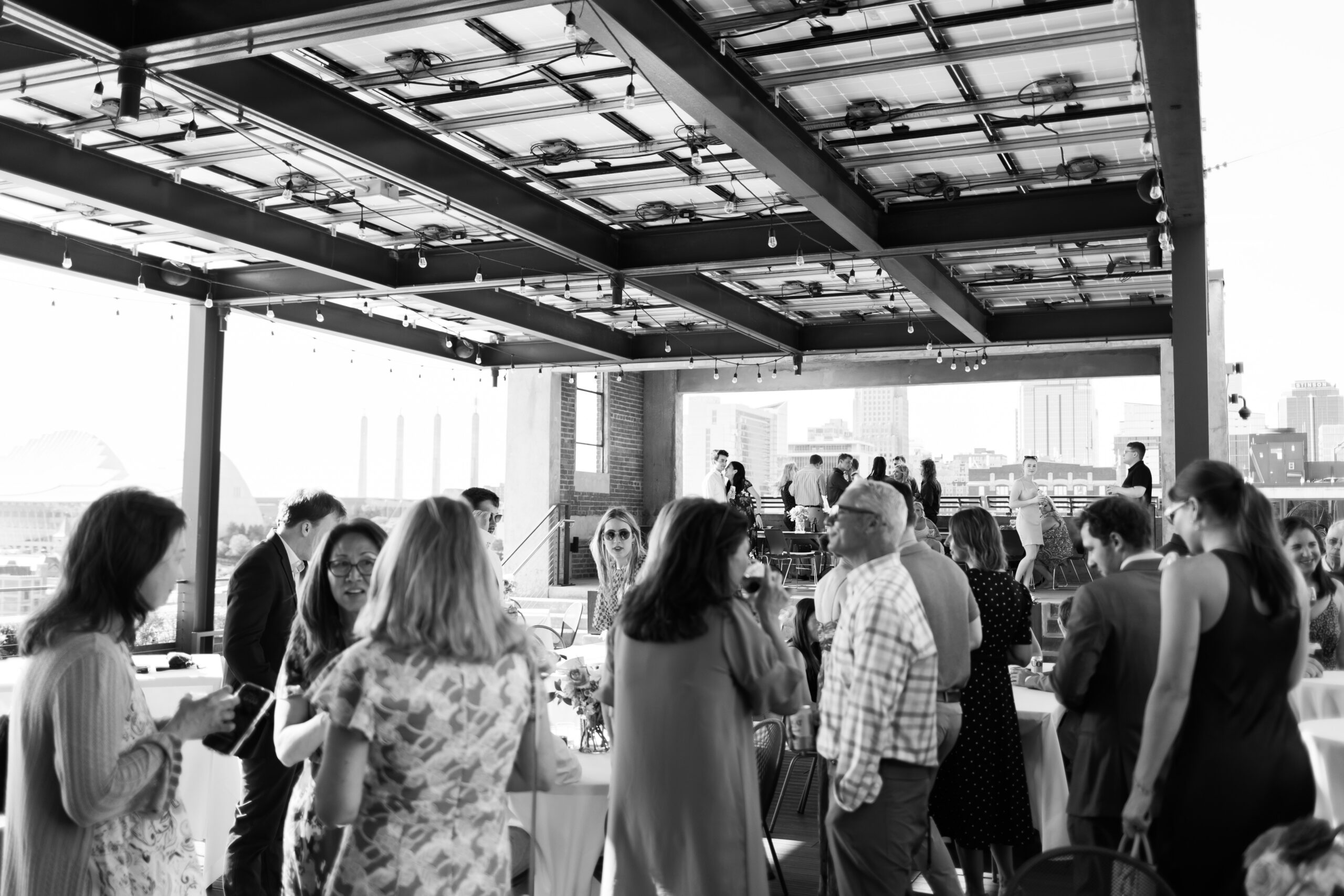 Wide view of a rooftop event space filled with guests mingling under string lights and a covered structure.