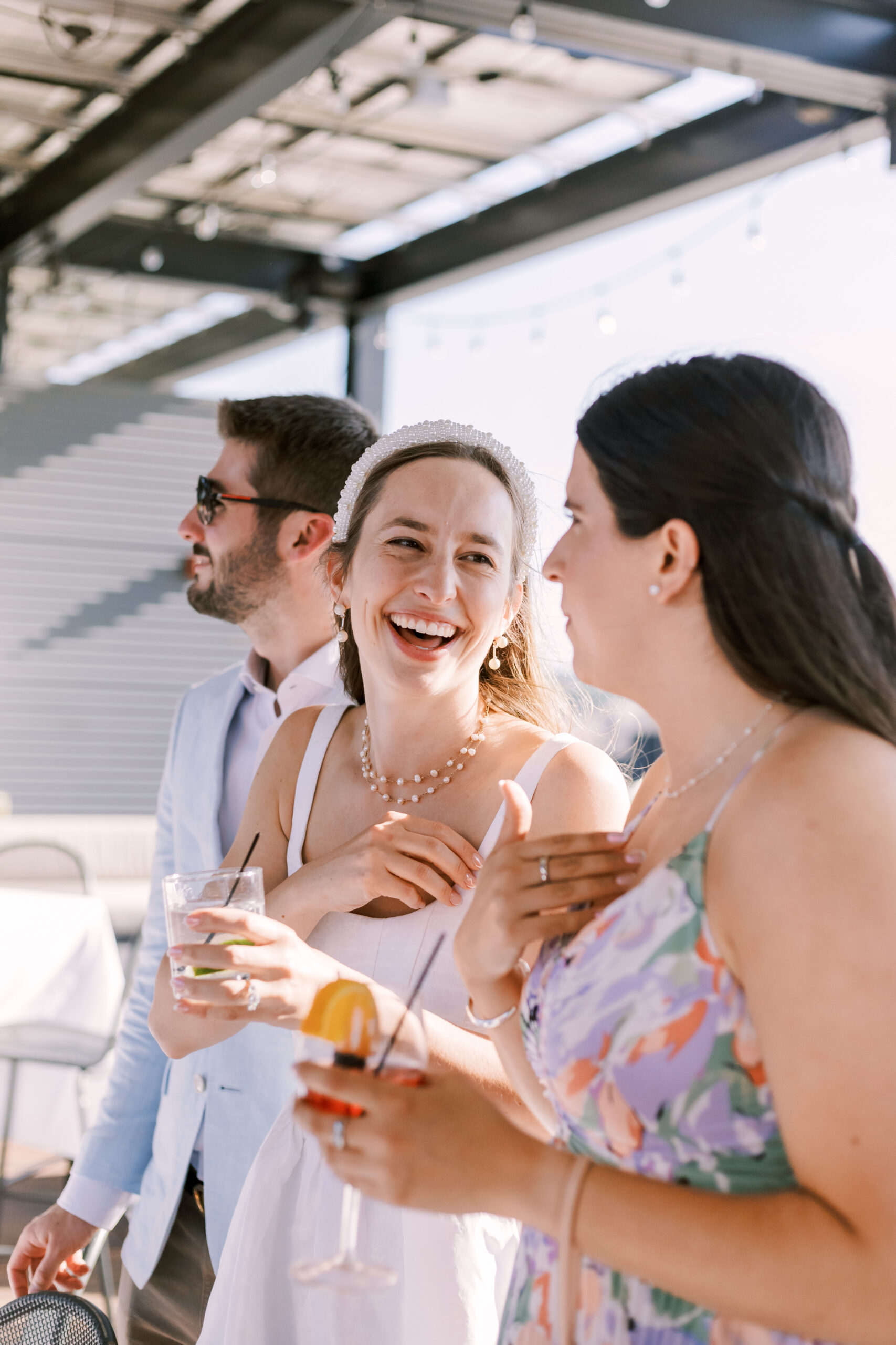 Close-up of guests laughing and talking while holding drinks at a sunlit rooftop gathering.