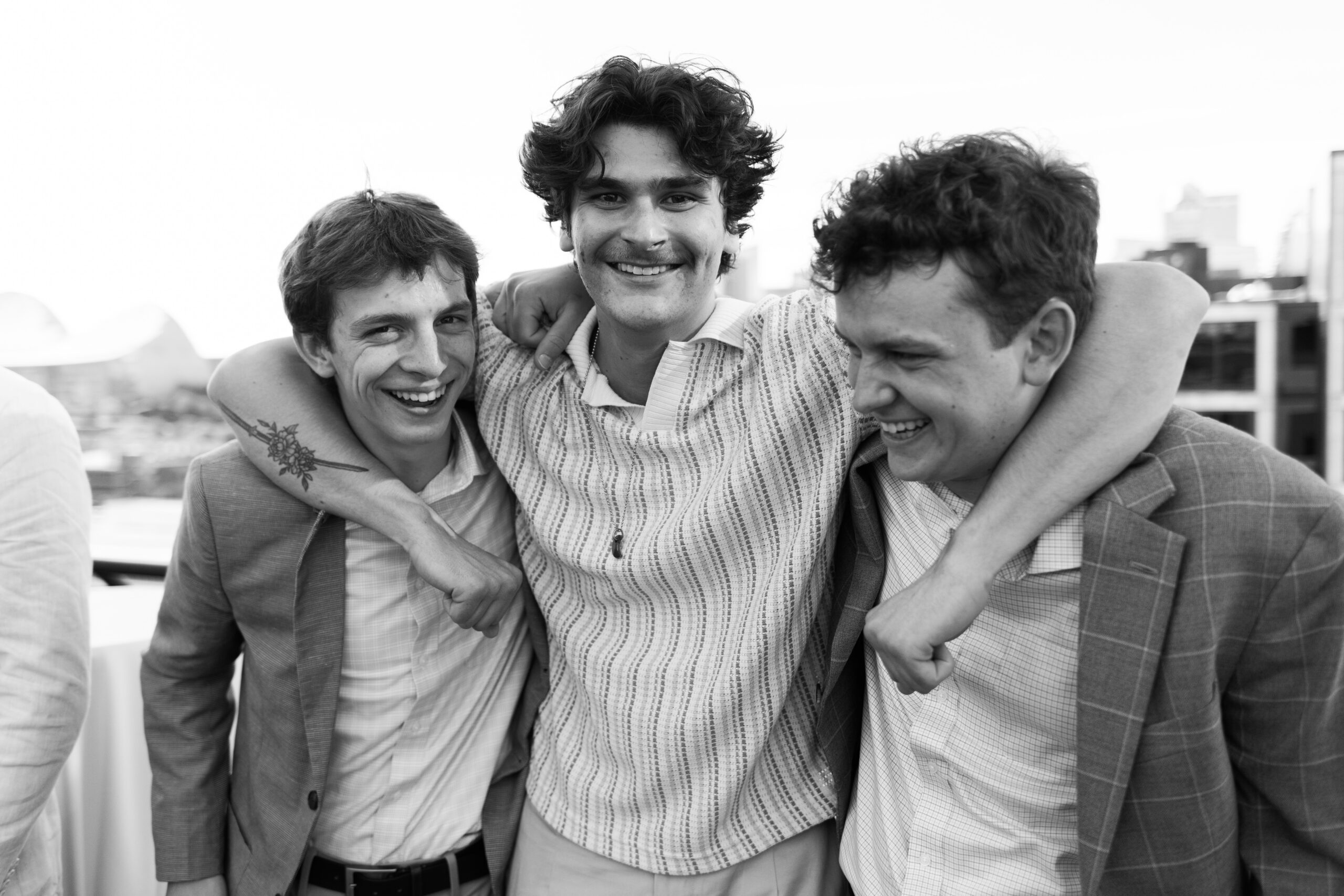 Three men in suits stand close together with arms around each other, smiling during a rooftop event.
