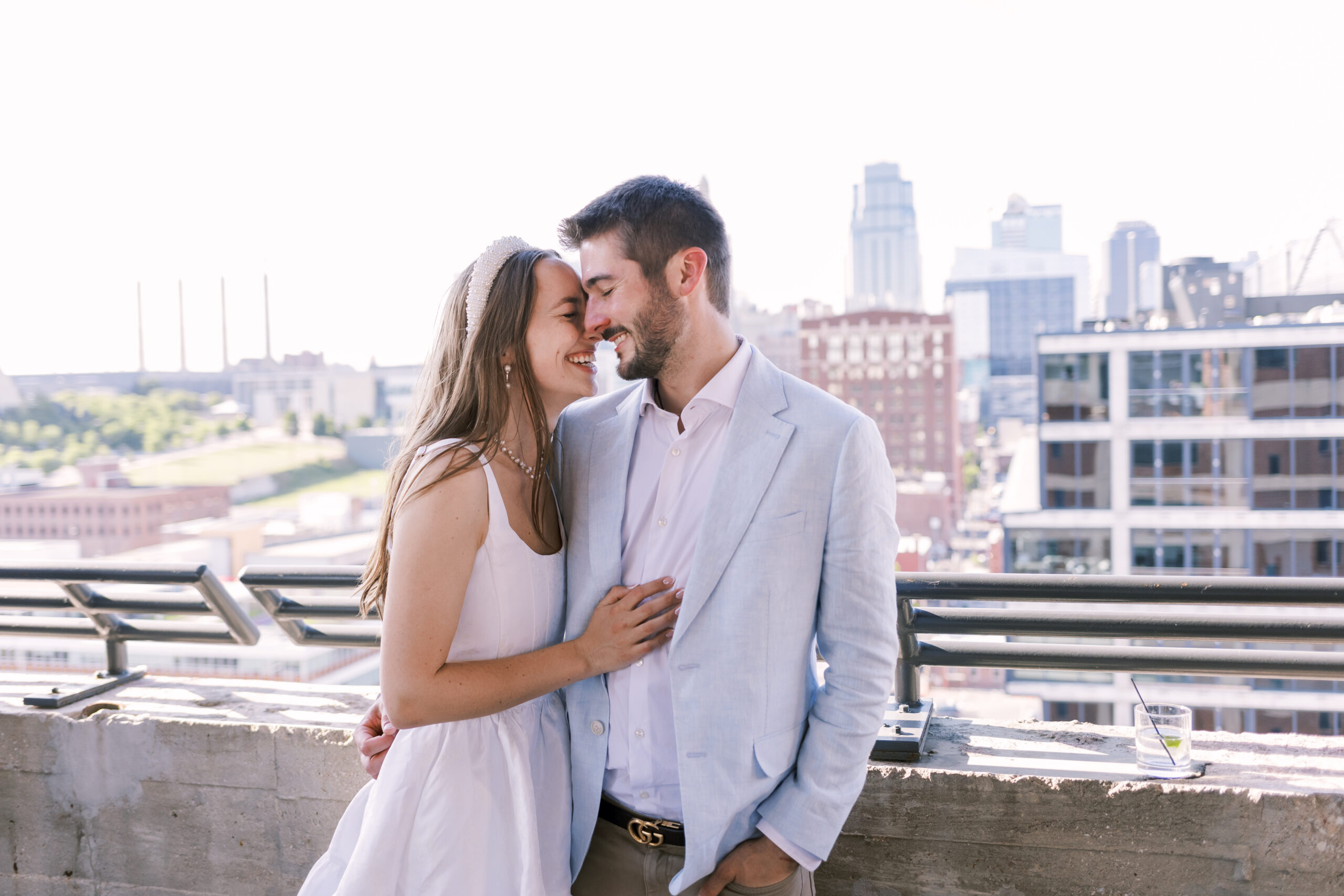 Couple stands close together on a rooftop terrace with a city skyline in the background.