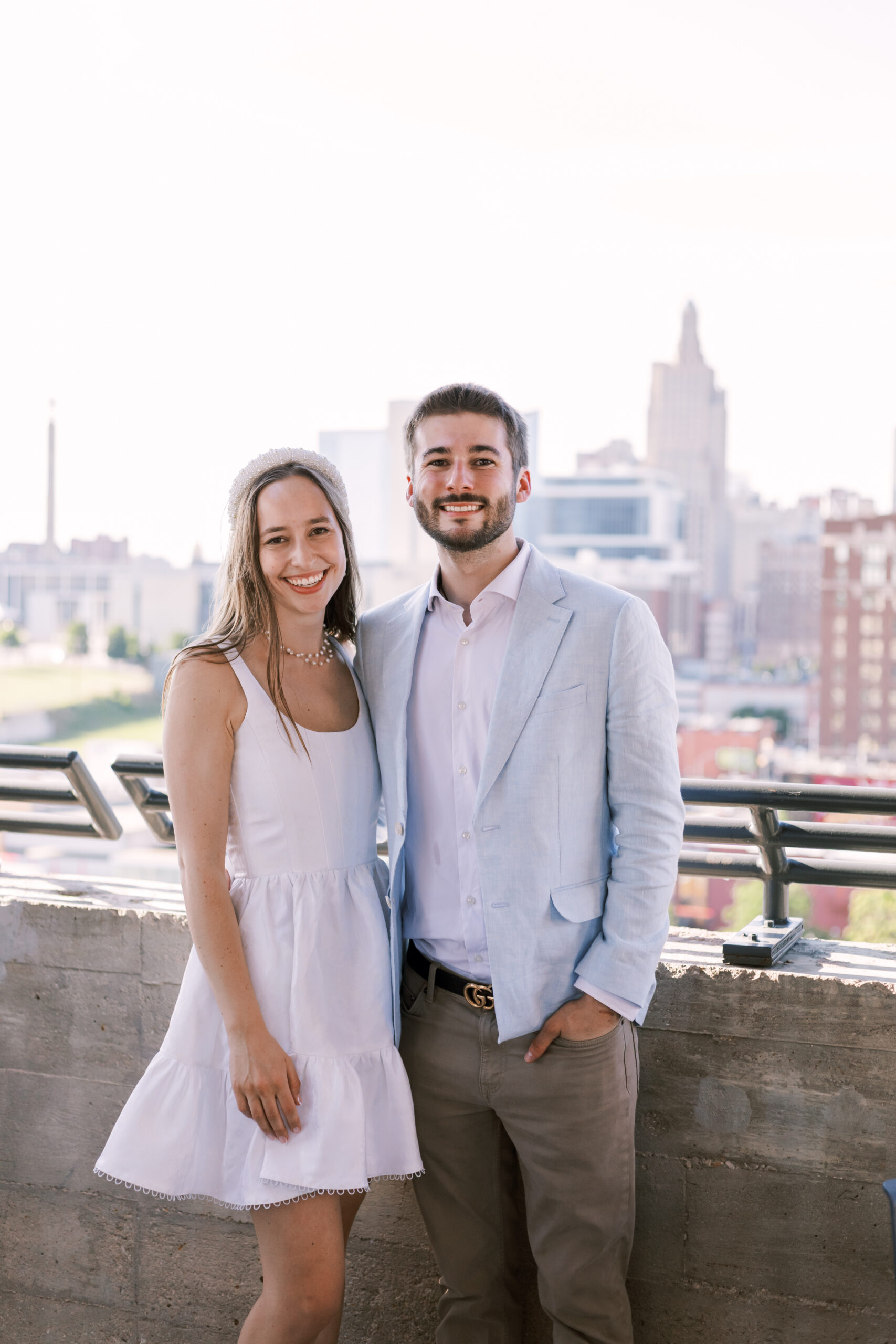 Portrait of a couple standing side by side, smiling at the camera on a rooftop with city views.