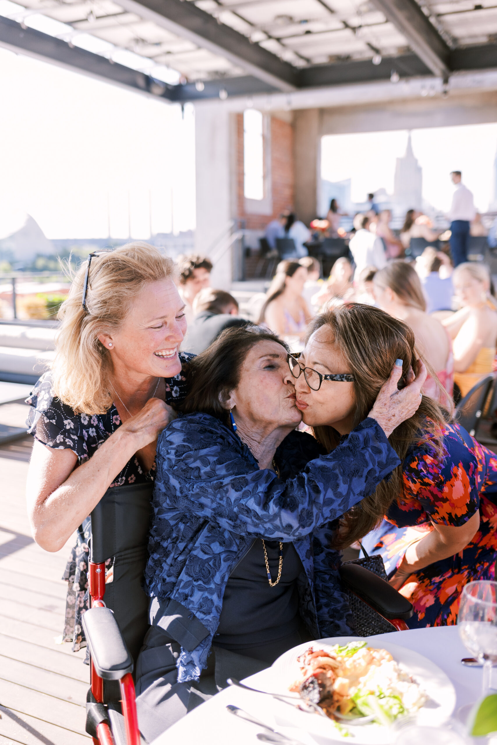 Two women greet and kiss an older seated guest at a table during an outdoor dinner reception.