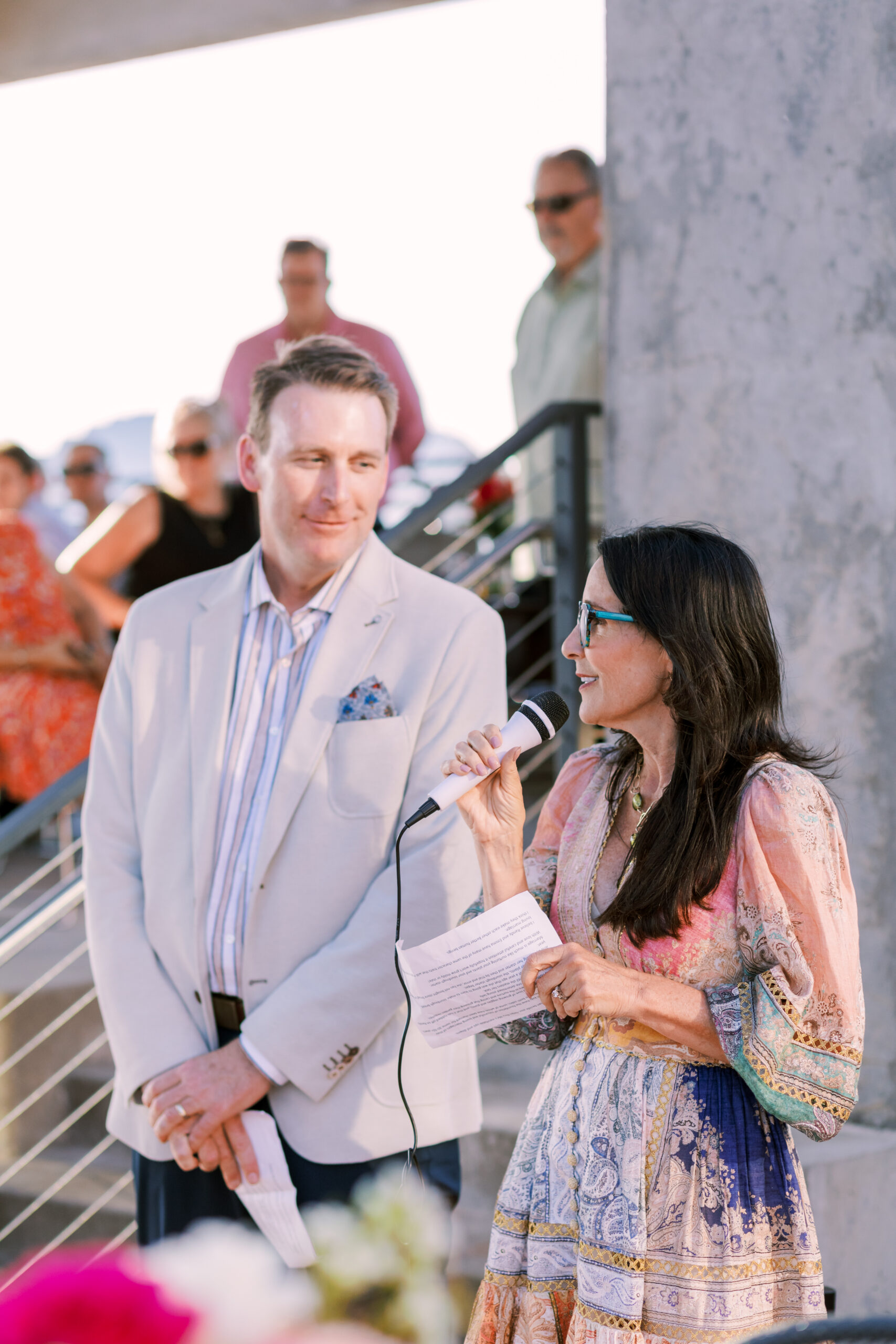Woman holding a microphone reads from a paper while speaking beside a man at an outdoor event.