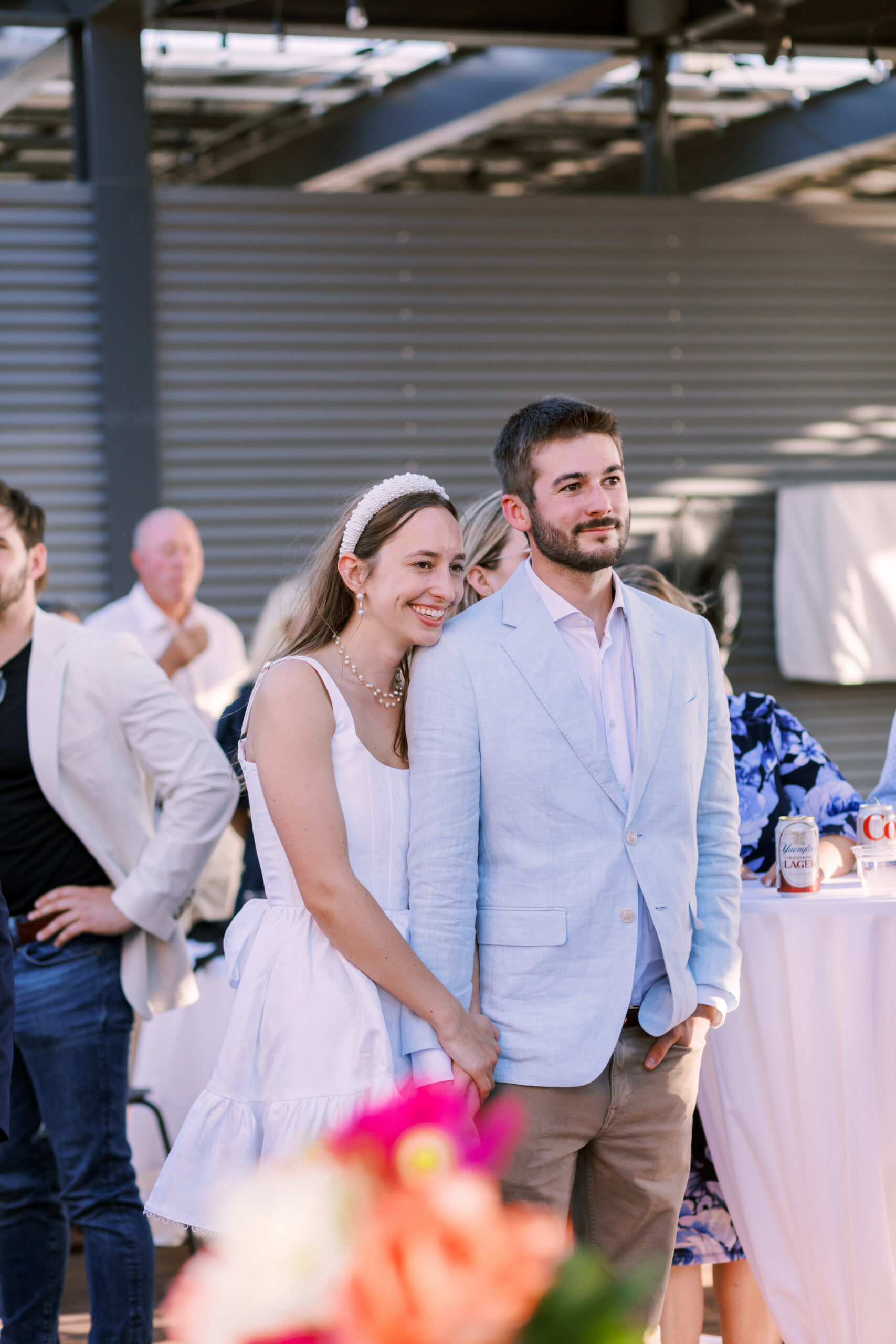 Couple stands together listening during speeches, surrounded by guests at a rooftop rehearsal dinner.