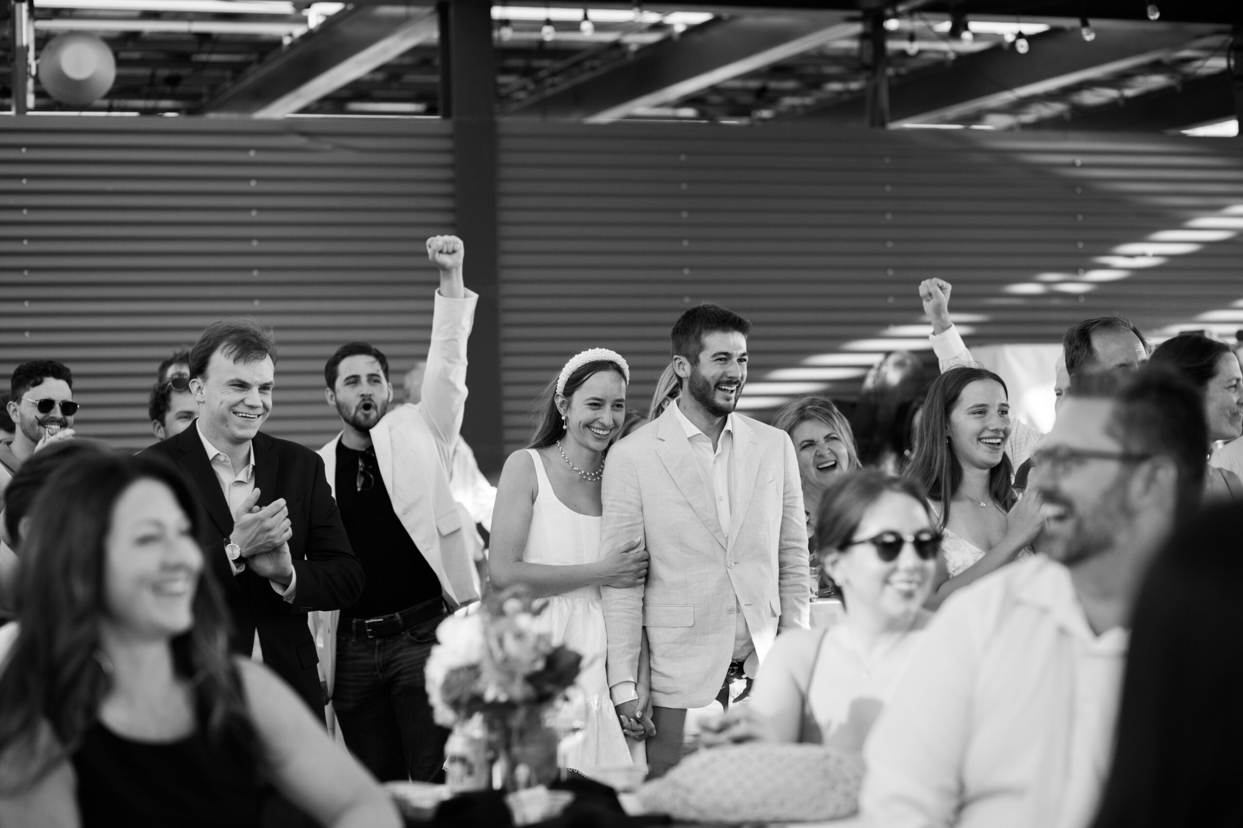 Guests cheer and raise hands while watching a couple during a lively reception moment.