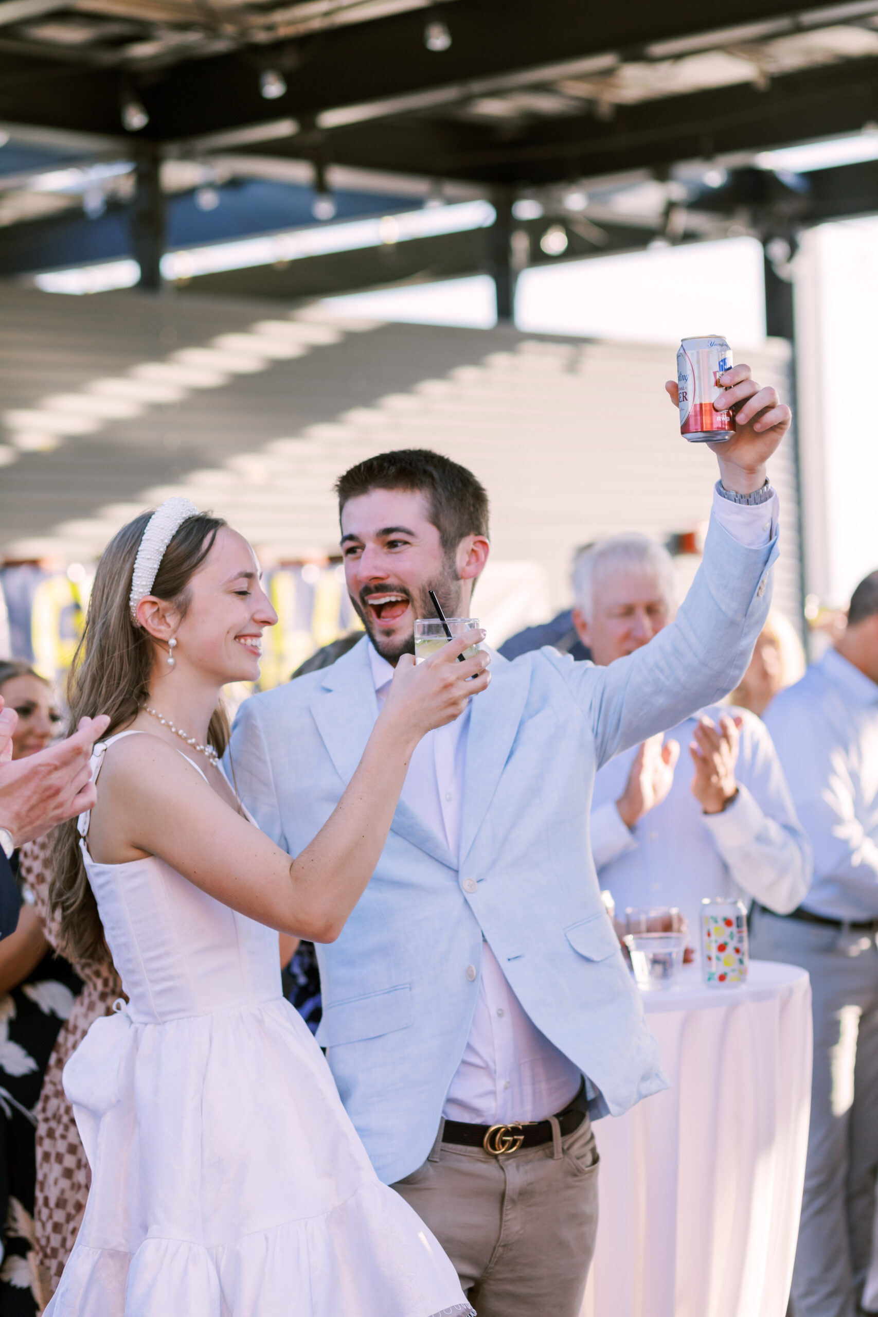 Couple raises drinks and smiles while guests applaud around them on a rooftop patio.