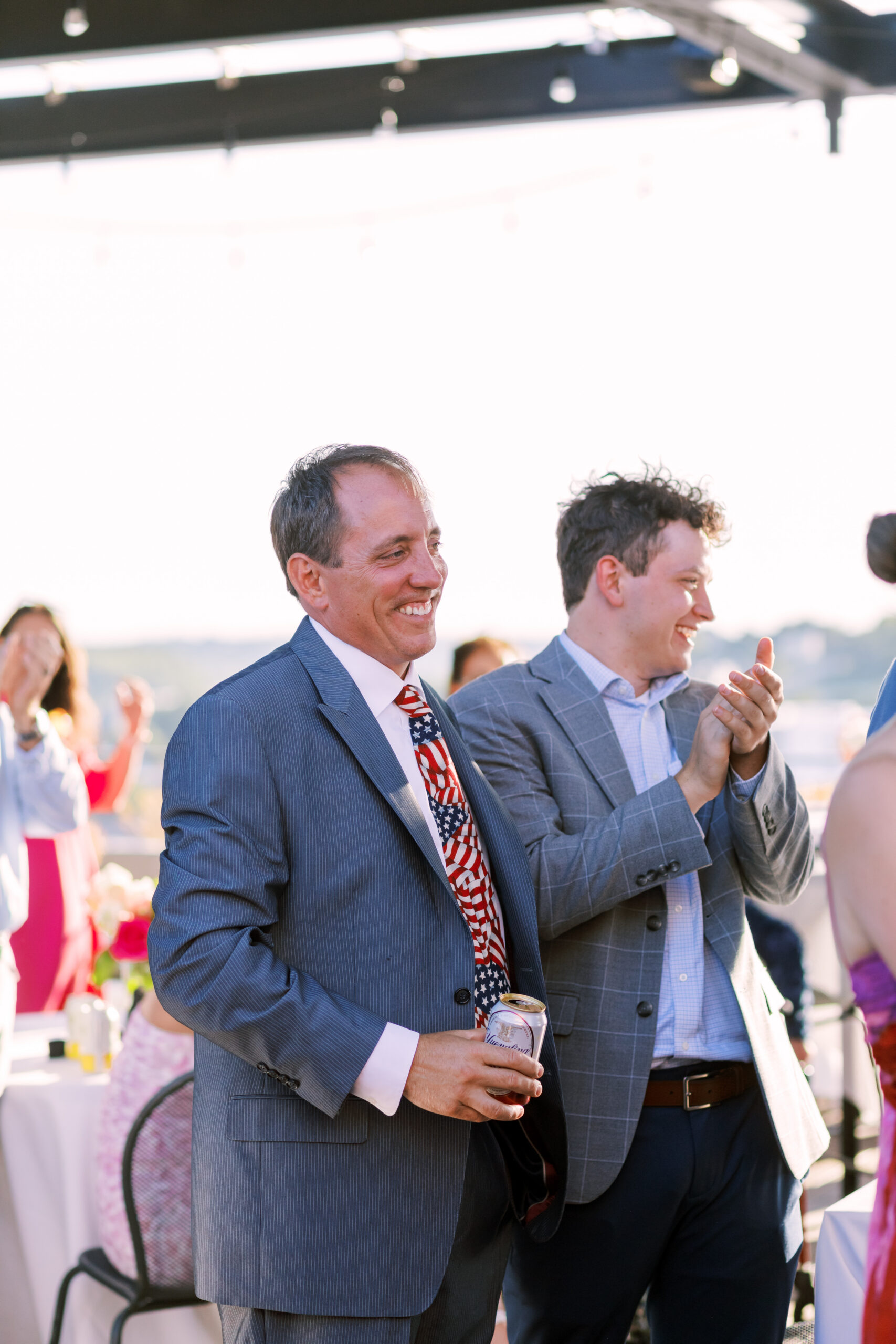 Two men in suits stand smiling and clapping during a speech at an outdoor evening gathering.