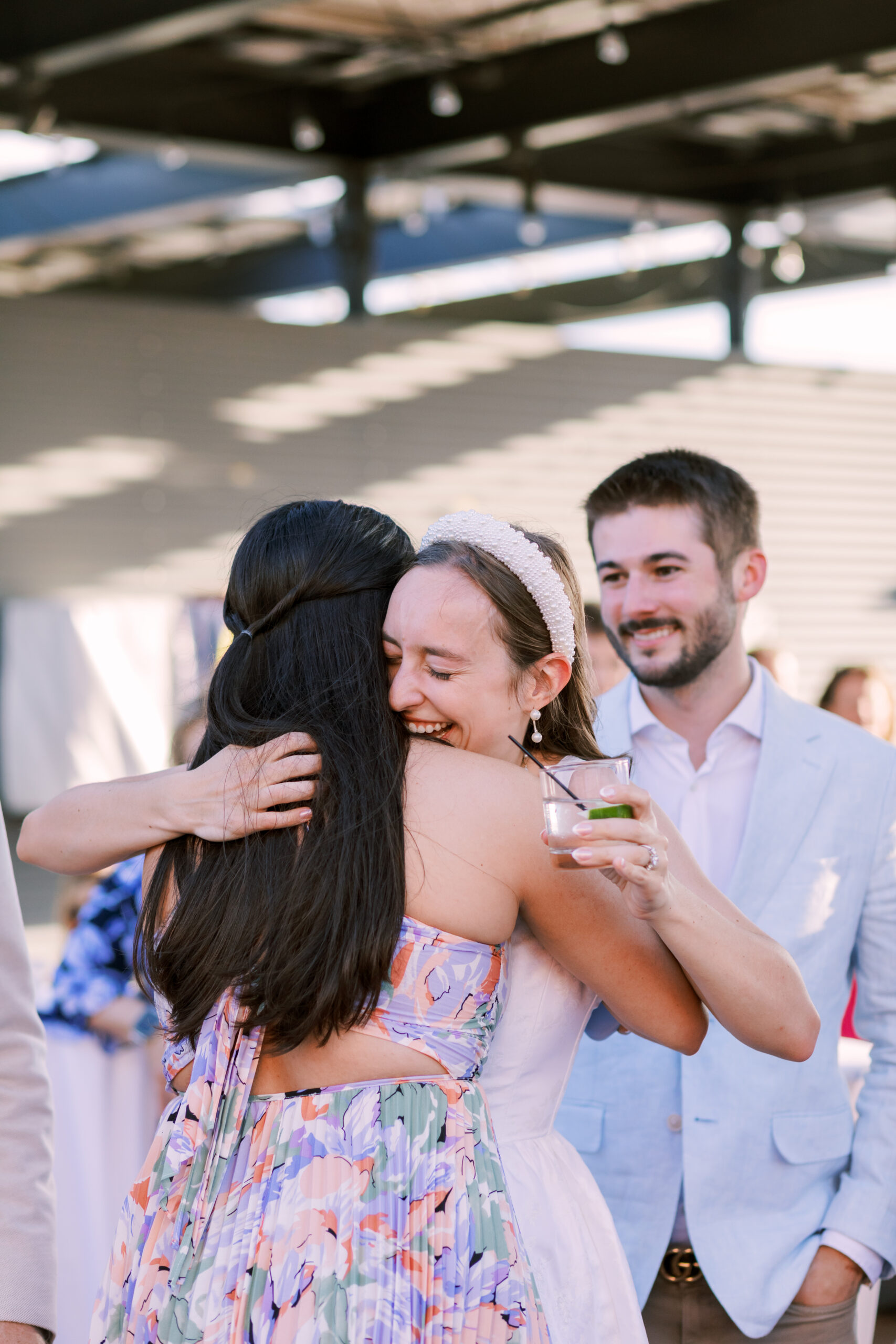 Two women embrace while holding drinks as a man stands nearby during a rooftop celebration.