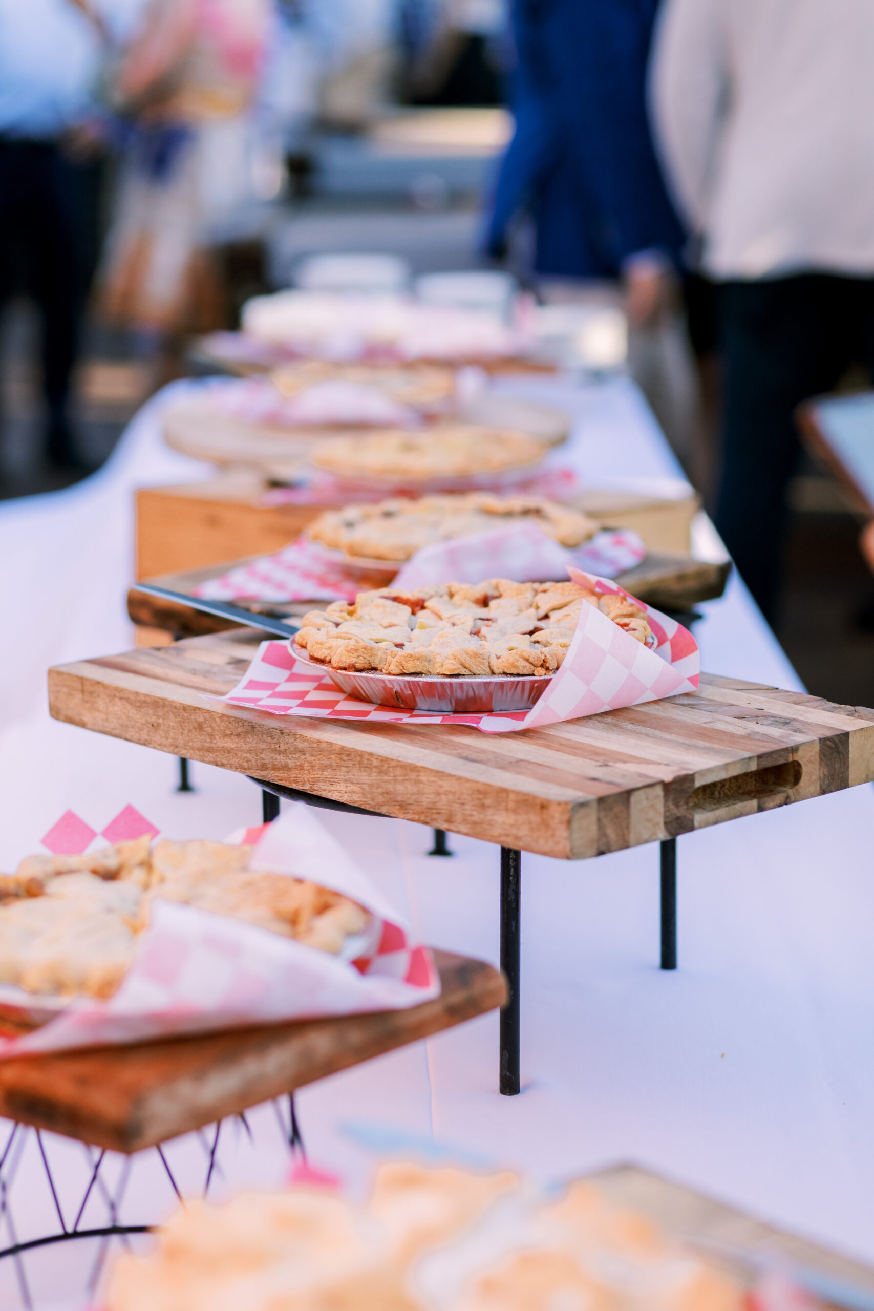 Row of assorted pies arranged on elevated wooden stands along a dessert table at an outdoor event.