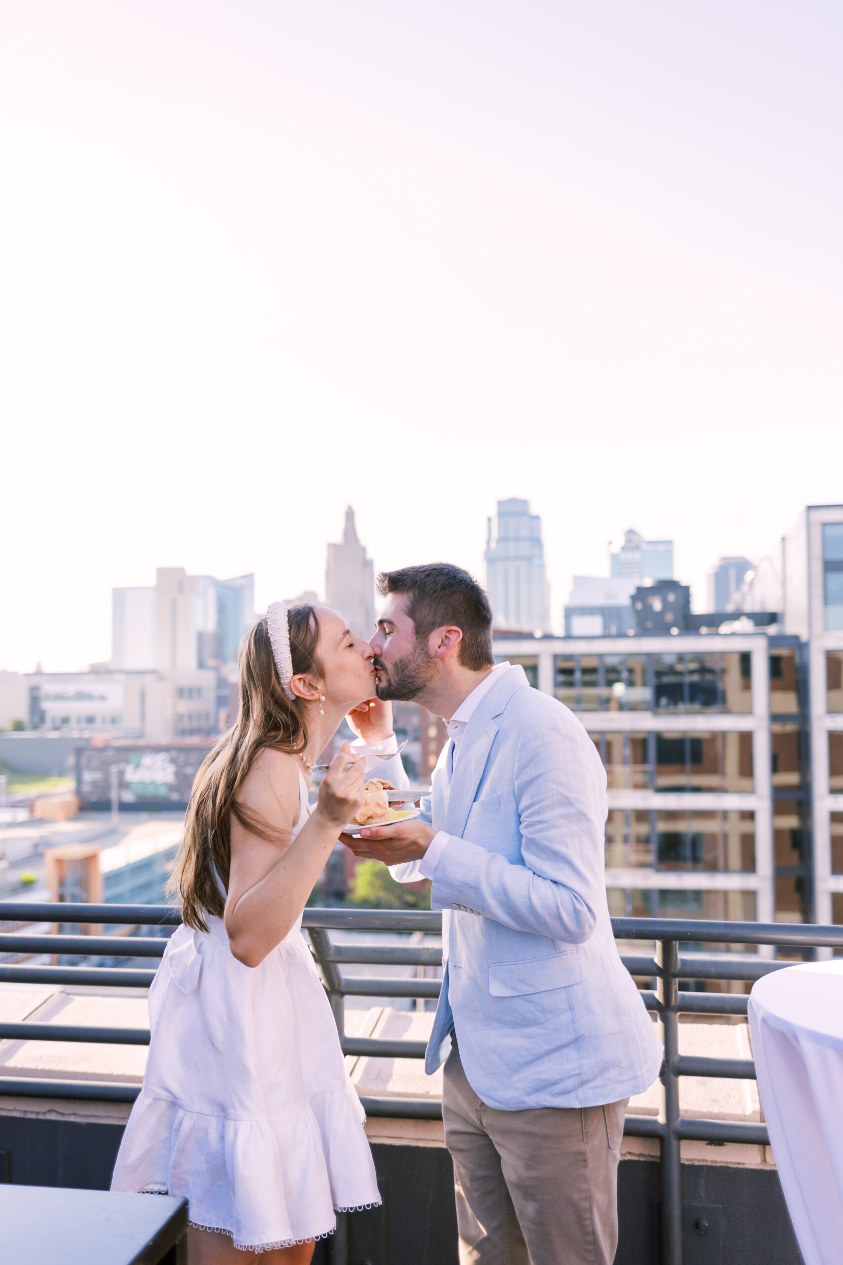 Couple stands on a rooftop terrace sharing a kiss while holding plates of pie with a city skyline behind them.