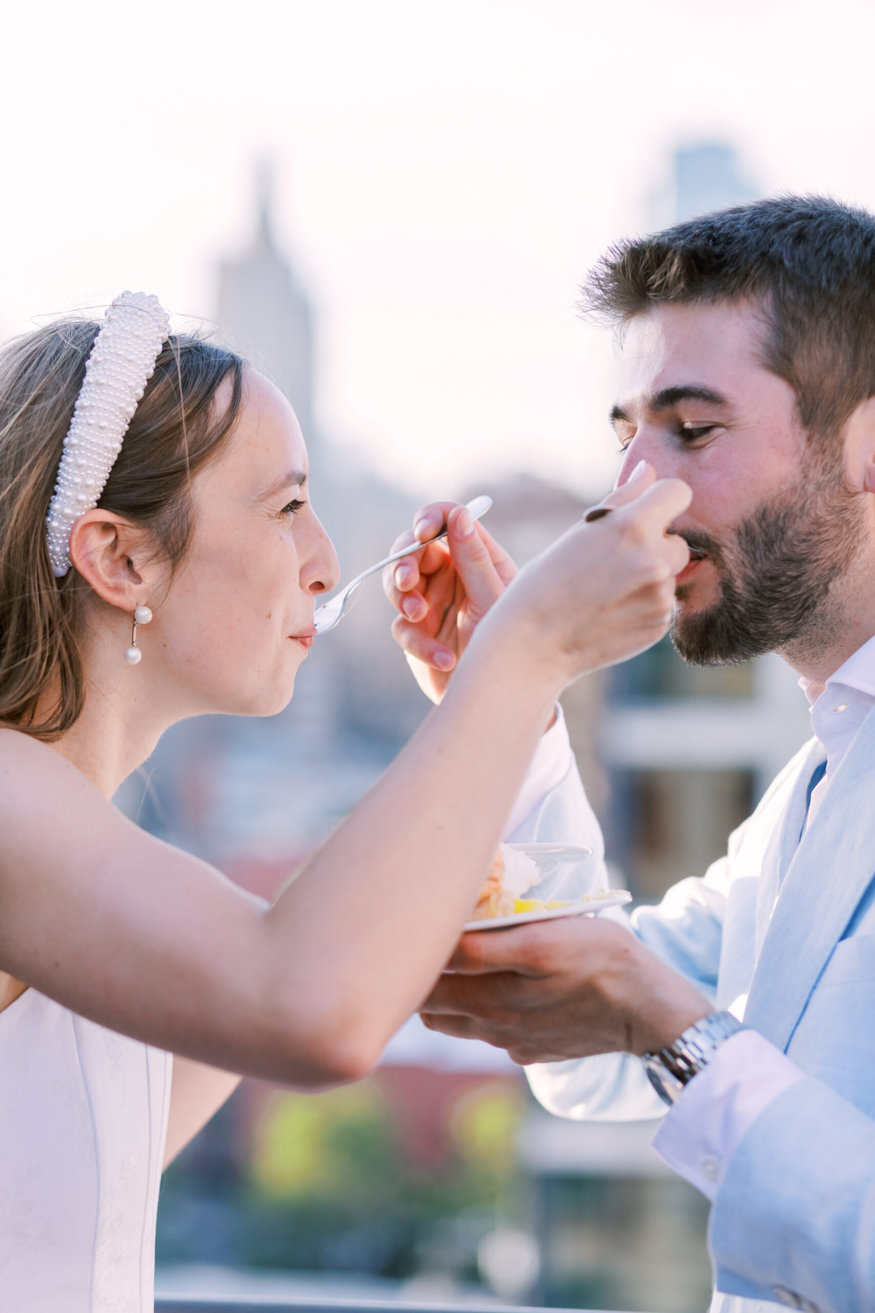 Bride feeds a bite of pie to her partner on a rooftop with a softly blurred city skyline in the background.