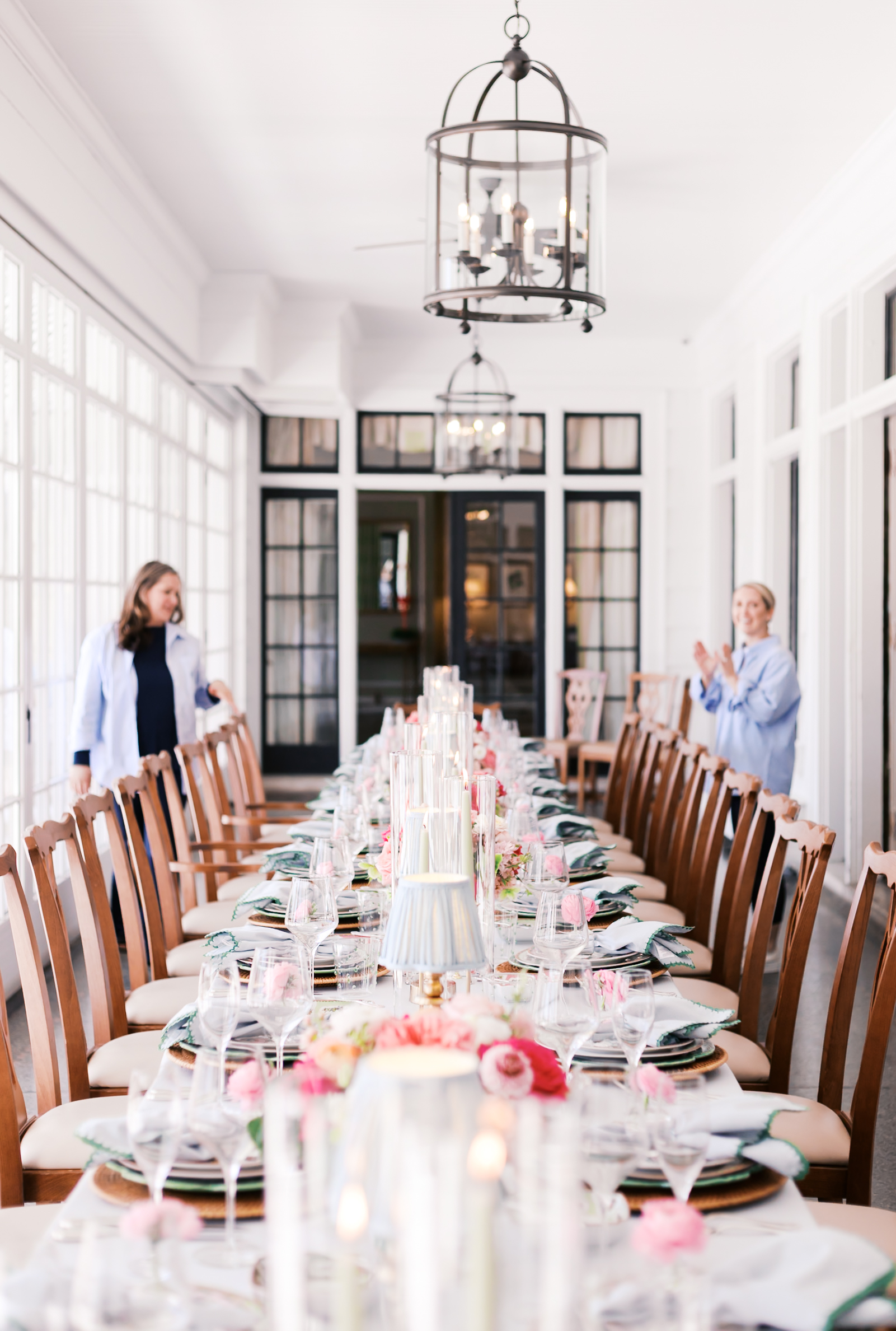 Long banquet table with floral arrangements and candles in bright white venue, two women adjusting settings.