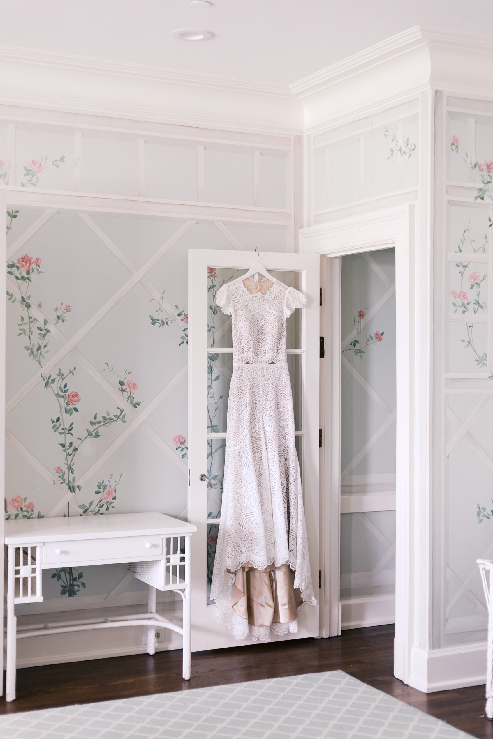 Lace wedding dress hanging on door in softly lit room with floral wallpaper and white furniture.