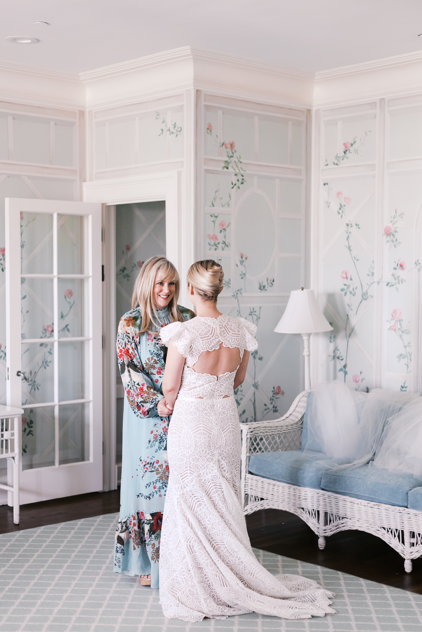 Bride in lace gown stands with woman in floral dress in bright, pastel-toned room.
