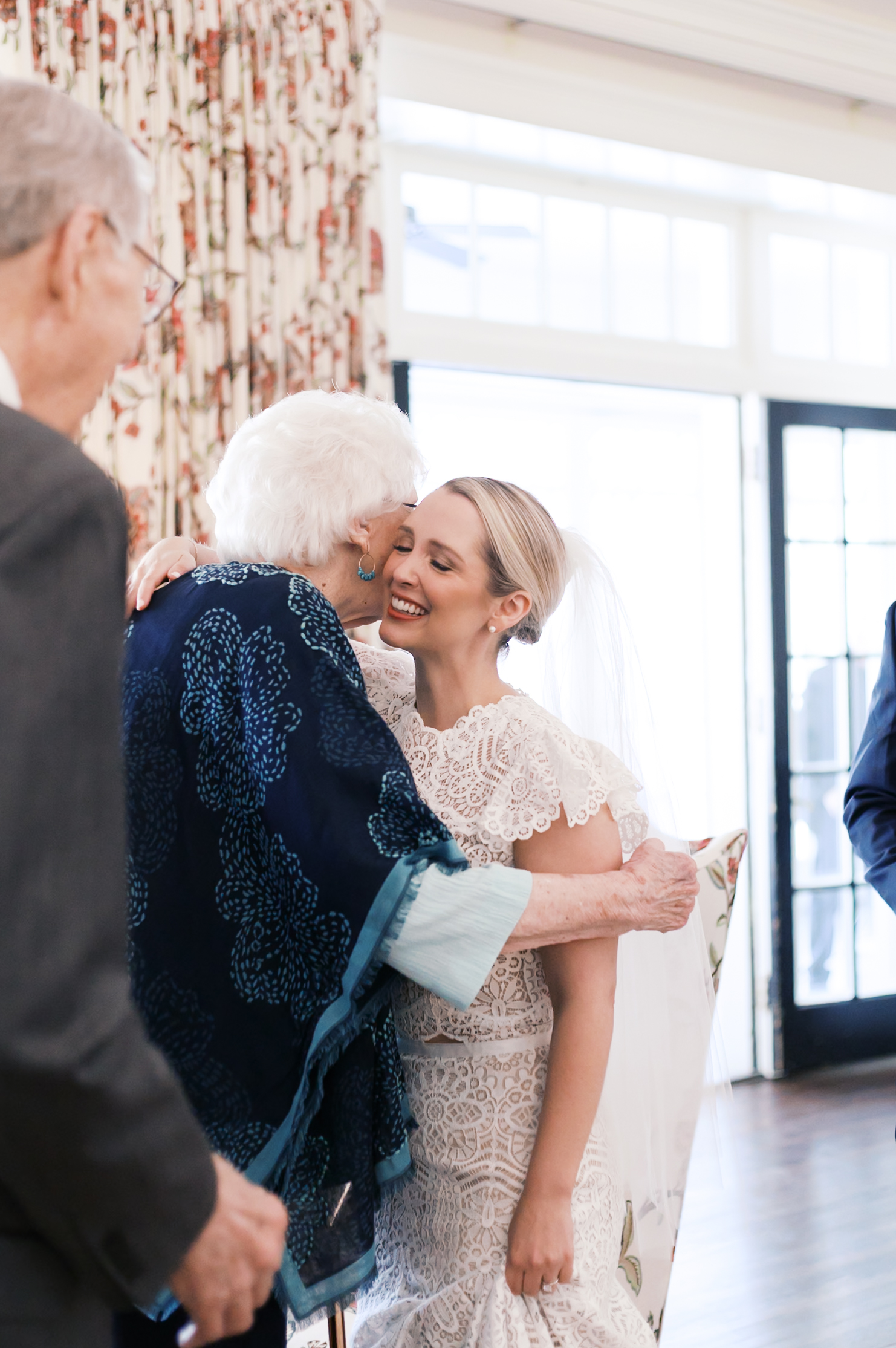 Bride hugs elderly woman while greeting guests at indoor wedding reception.